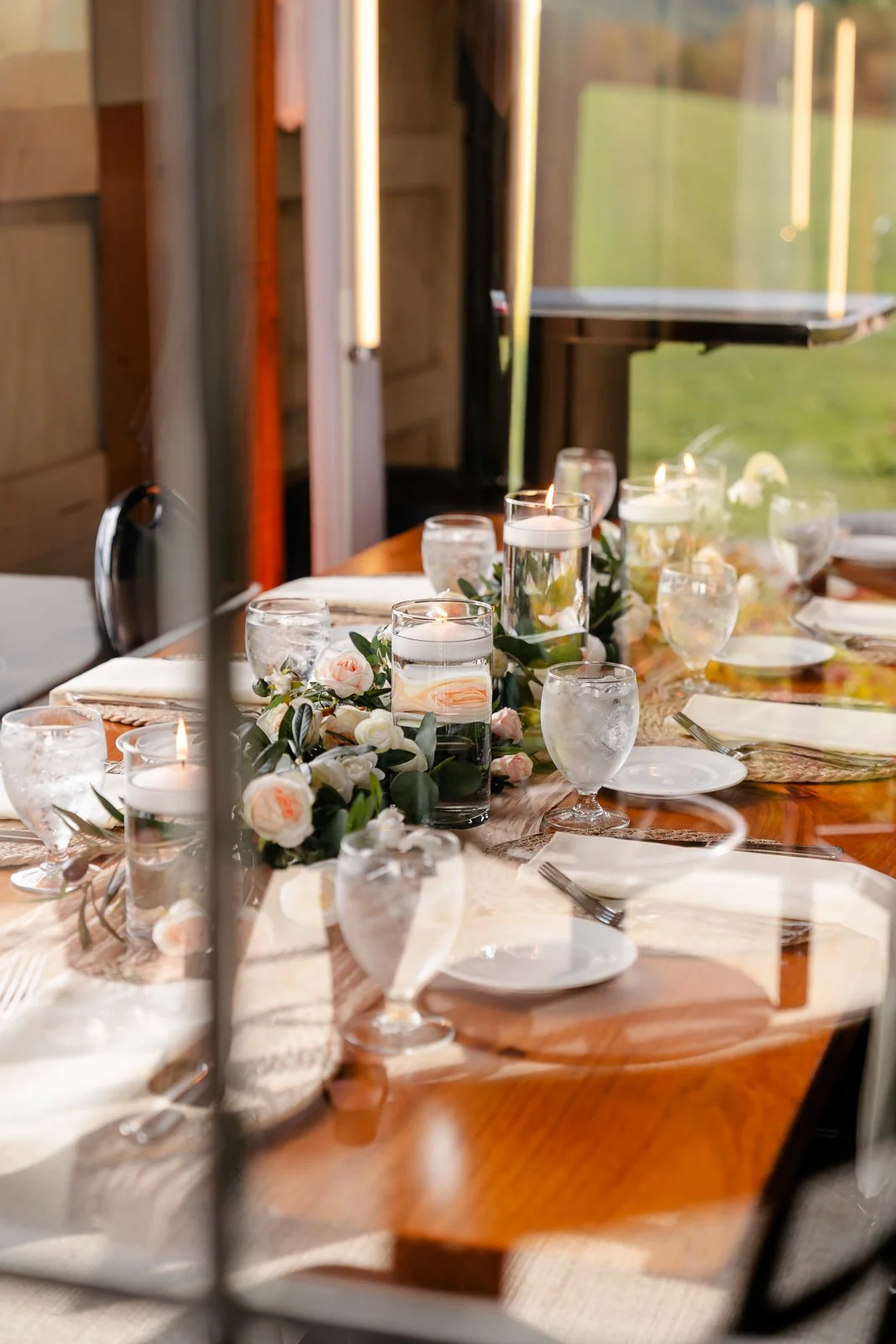 Elegant table setting with white flowers, candles, glassware, and plates in a sunlit room with wooden walls.