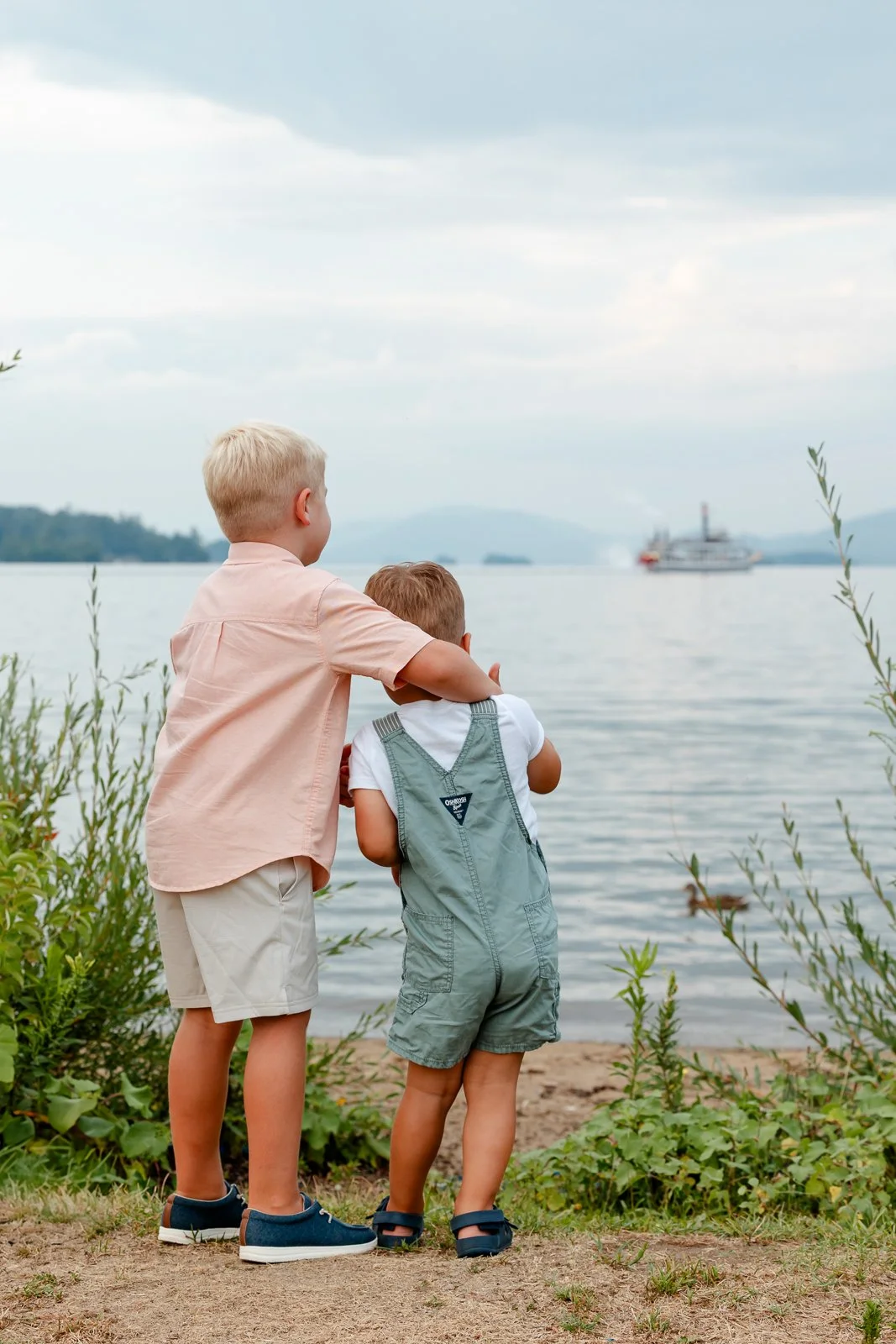 Two young boys stand on the shore, looking out over a body of water with a boat in the distance, surrounded by greenery and a cloudy sky.