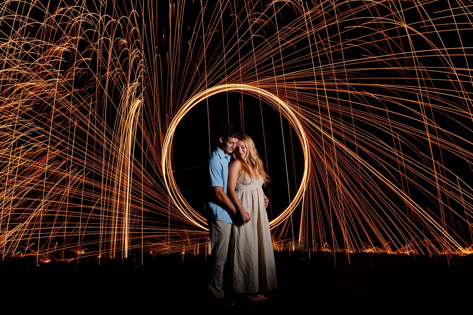 A couple standing close together, smiling, in front of a light painting with swirling sparks forming a heart shape in the dark.