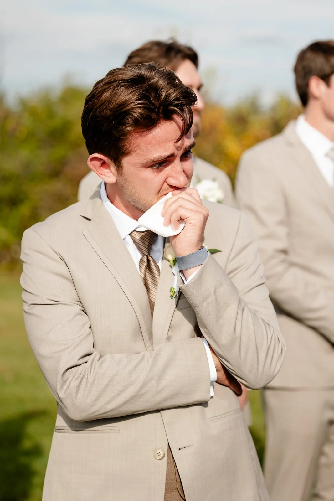 A young man in a beige suit appears emotional, holding a tissue to his face during an outdoor ceremony with other men in similar suits in the background.