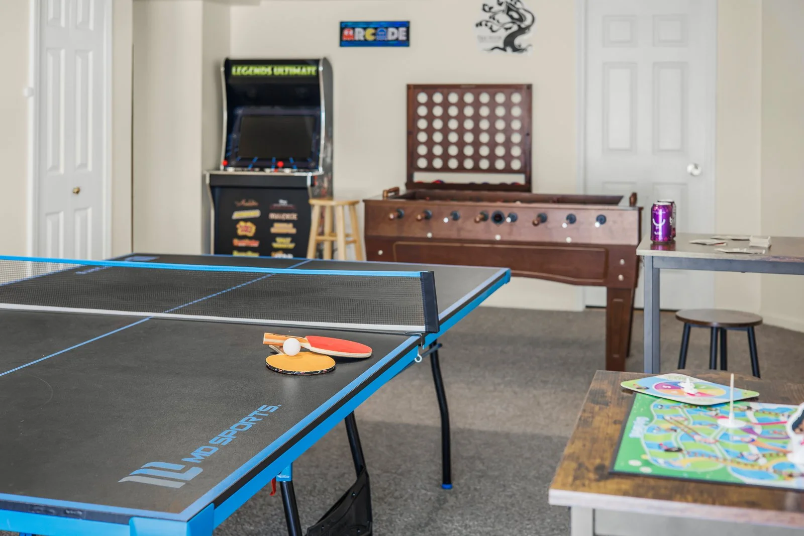 An indoor game room with a ping pong table, arcade game, foosball table, and board games on a wooden table.
