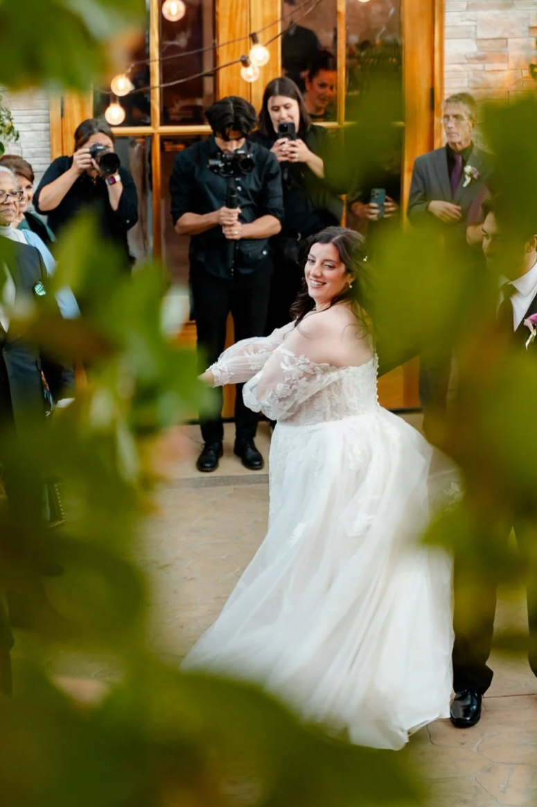 A bride in a white lace wedding dress smiling during a wedding ceremony, surrounded by guests and photographers in a warmly lit indoor venue with string lights.