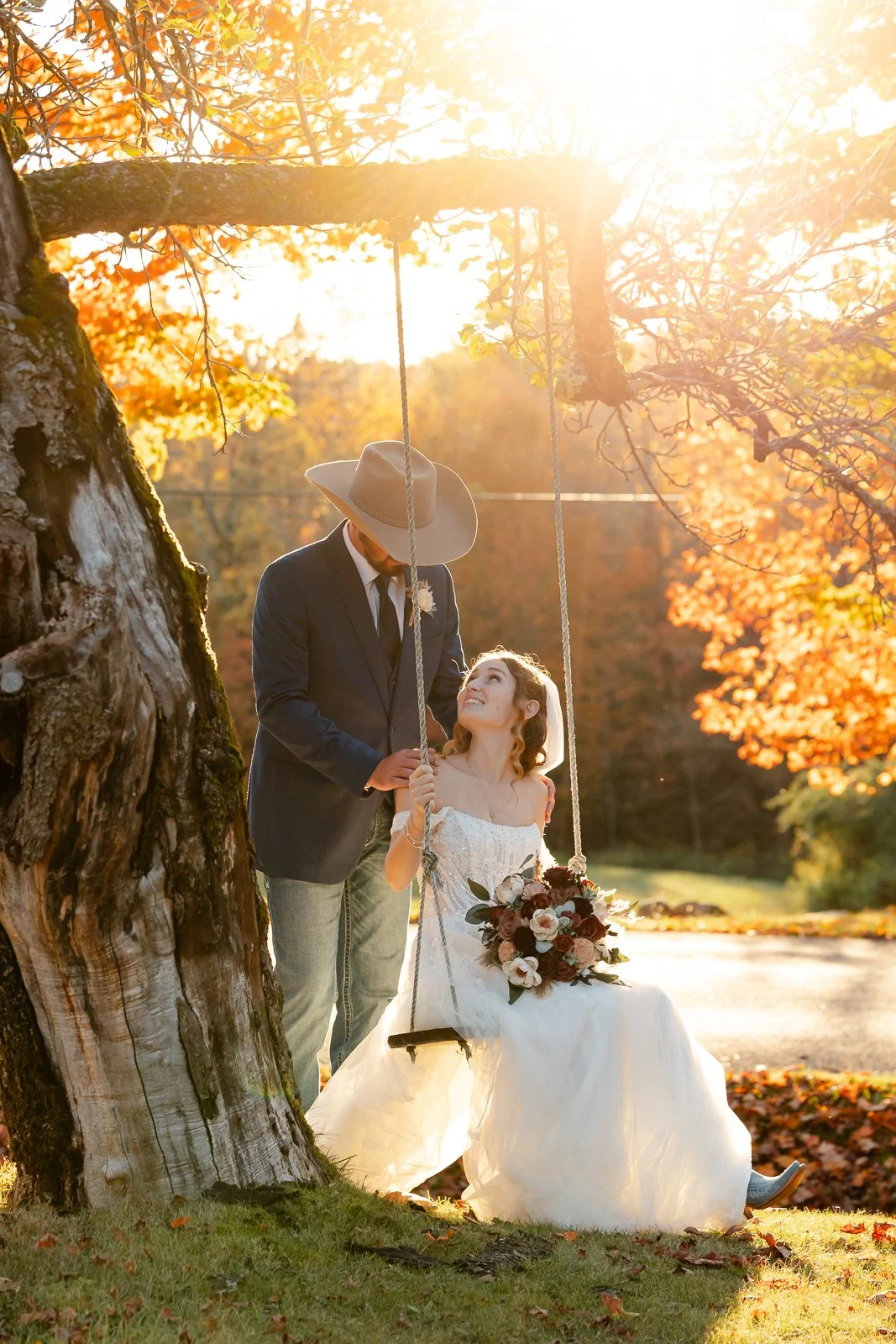 A bride and groom sharing a moment on a swing hanging from a tree in an autumn setting, with warm sunlight filtering through orange and yellow leaves, during a wedding photoshoot.