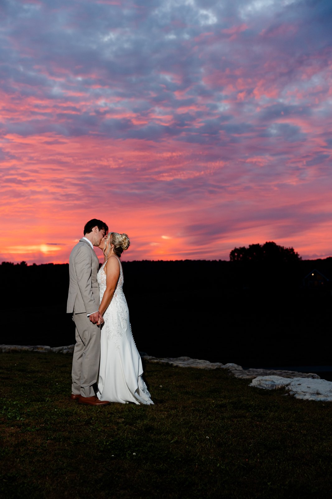 A bride and groom kissing during sunset on a grassy area with trees in the background.