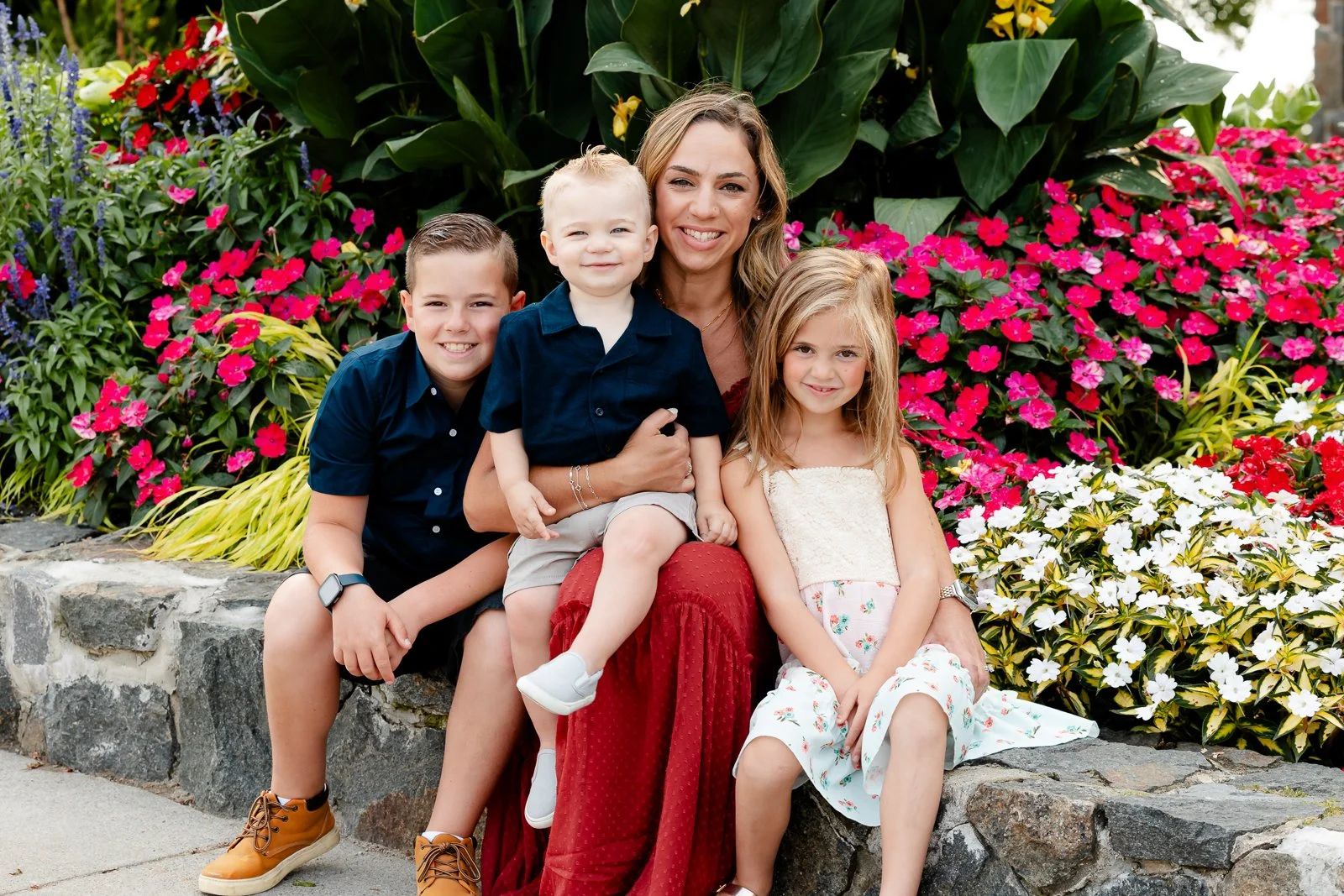 A woman and three children sitting on a stone ledge in front of colorful flowers, smiling at the camera.