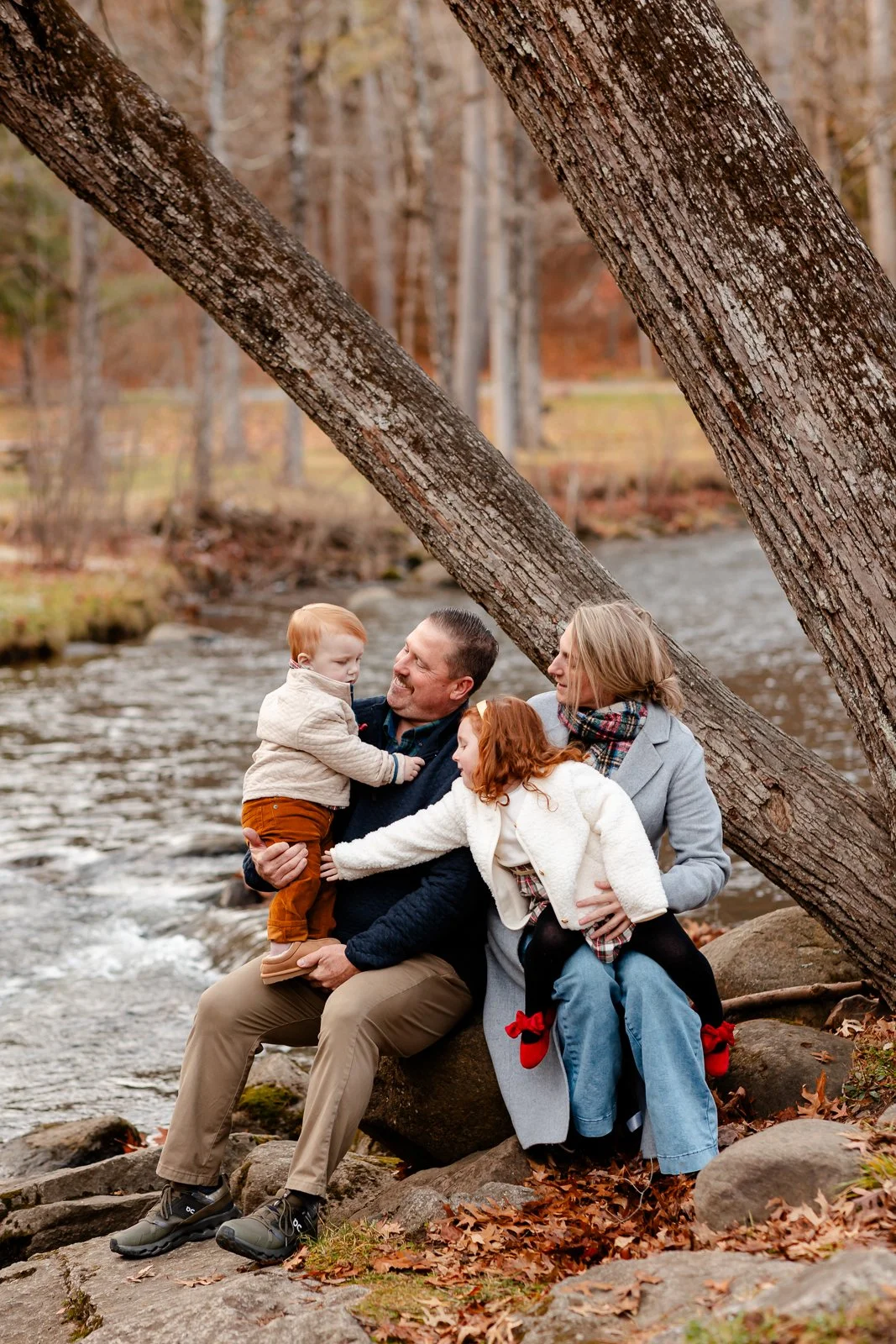 A family of four sitting on rocks by a creek in autumn. The father is holding a young boy, while the mother and daughter are interacting with the boy and smiling.