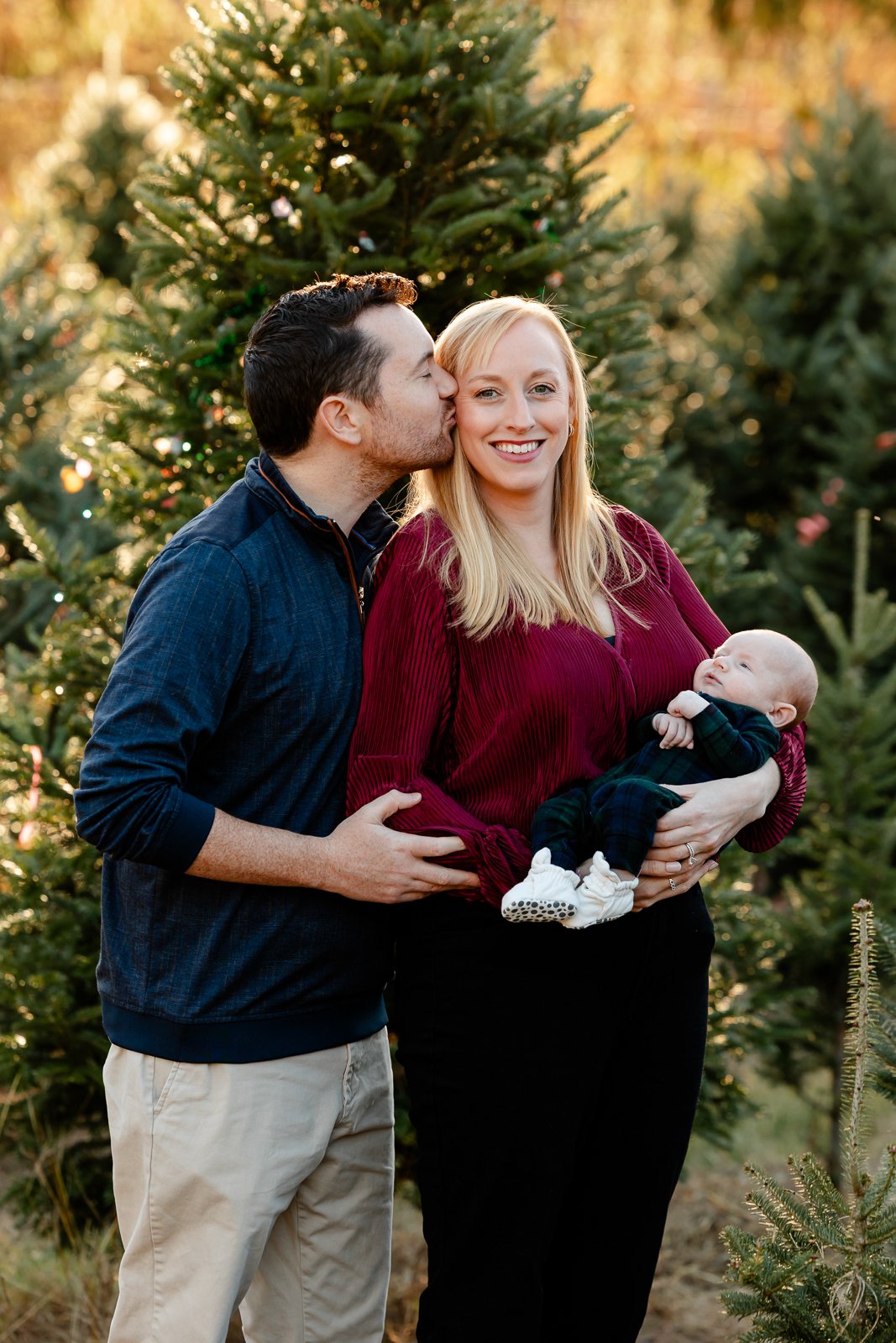 A family of three, including a mother, father, and infant, stands outdoors among Christmas trees. The father kisses the mother on the cheek, and she smiles while holding the baby. The scene is set in a natural, festive environment with warm sunlight.