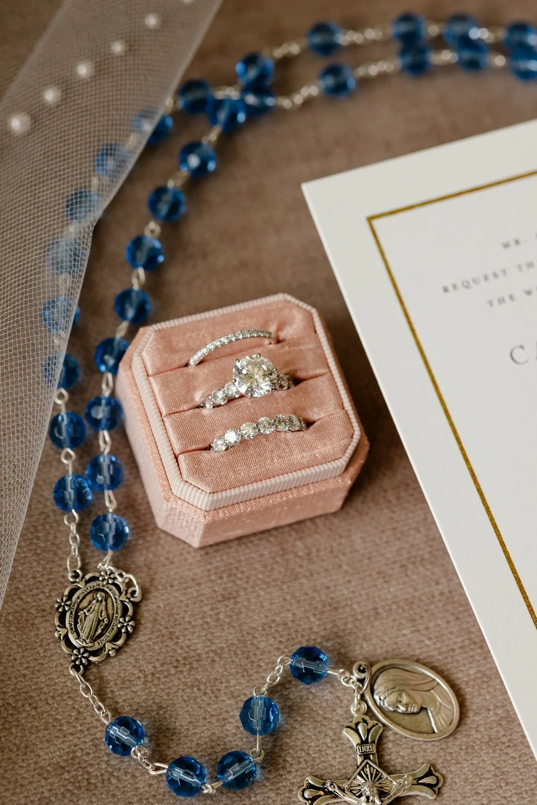 Close-up of a pink velvet ring box containing three diamond rings and a gold bracelet with blue beads and religious pendants, on a brown fabric surface with part of a white and gold wedding invitation.