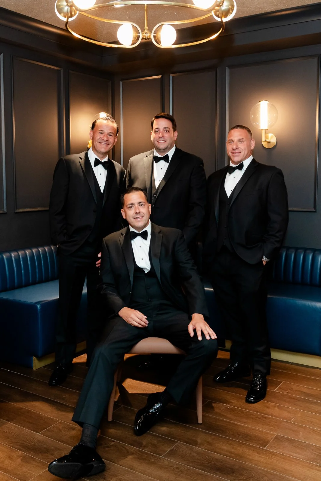 Five men in tuxedos posing in a dark room with blue seating and modern lighting.