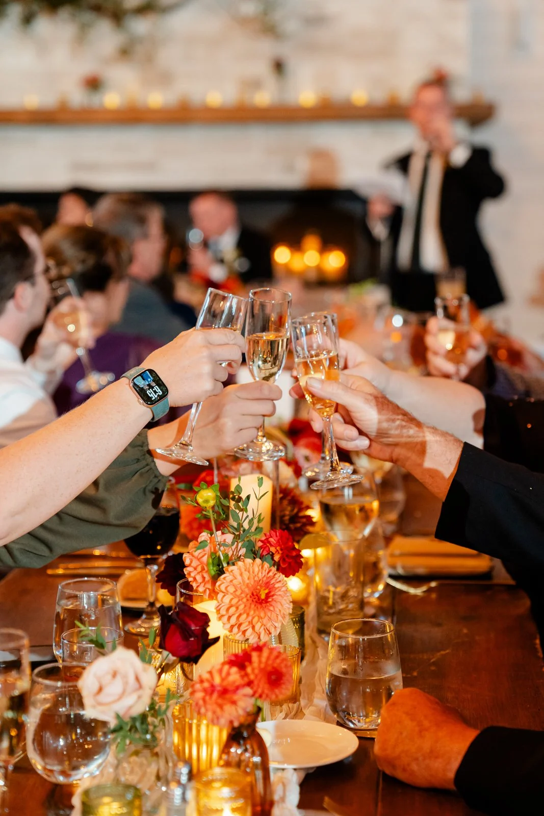 People raising glasses of champagne in a toast at a festive dinner party with floral decorations and a fireplace in the background.