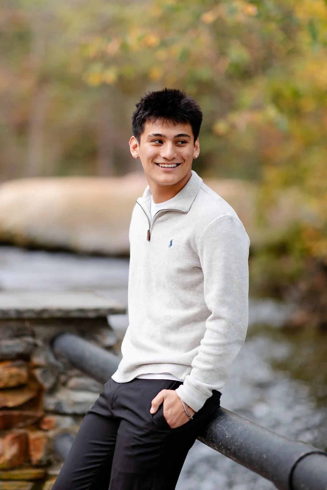 A young man with dark hair, wearing a light gray quarter-zip sweater and black pants, leaning against a black railing outdoors near a stream, smiling and looking to the side with autumn trees in the background.
