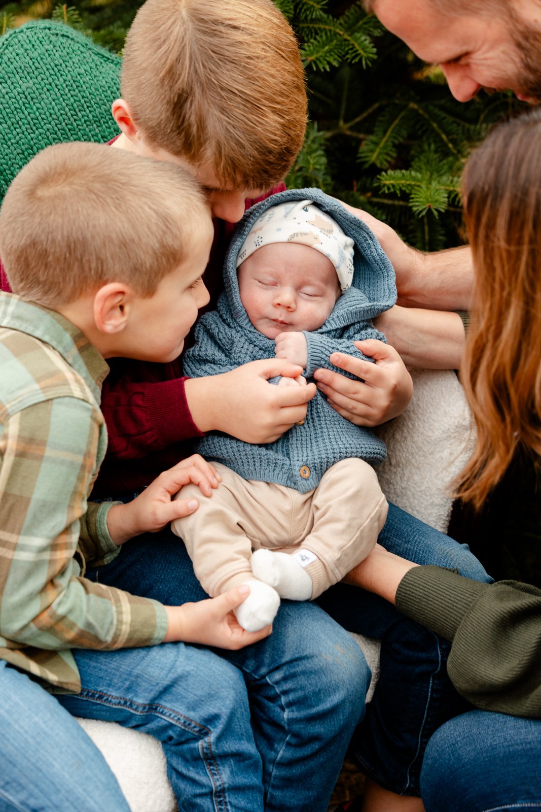 A group of family members, including two young boys, a man, and a woman, gathered around a sleeping baby dressed in a blue hoodie and beige pants, with green trees in the background.