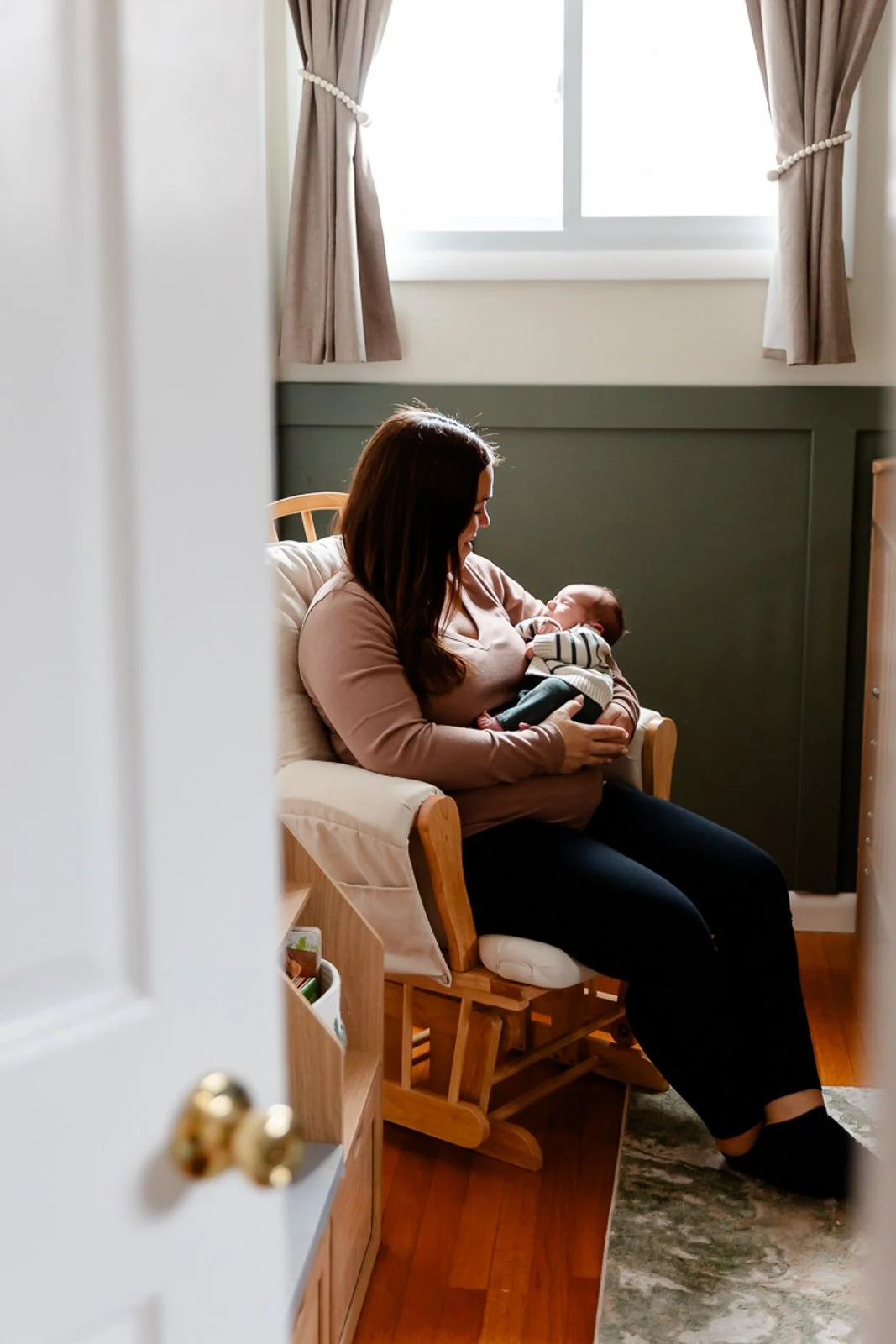 A woman sitting in a wooden chair, breastfeeding a baby in a cozy, softly lit room. The woman wears a beige top and black pants, with long brown hair. There is a window with beige curtains behind her, and a green wainscoting on the wall. A small rug is on the wooden floor.