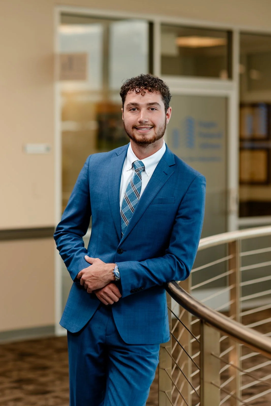 A young man in a blue suit, white shirt, and plaid tie standing indoors with his arms crossed, smiling at the camera.