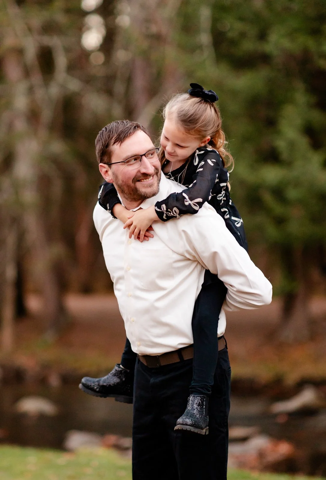 A man with glasses and a beard carrying a young girl on his shoulders, both smiling and looking at each other outdoors in a wooded area.