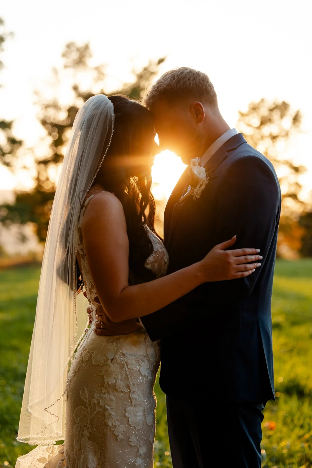 A bride and groom standing close with foreheads touching at sunset, outdoors in a grassy area with trees in the background.
