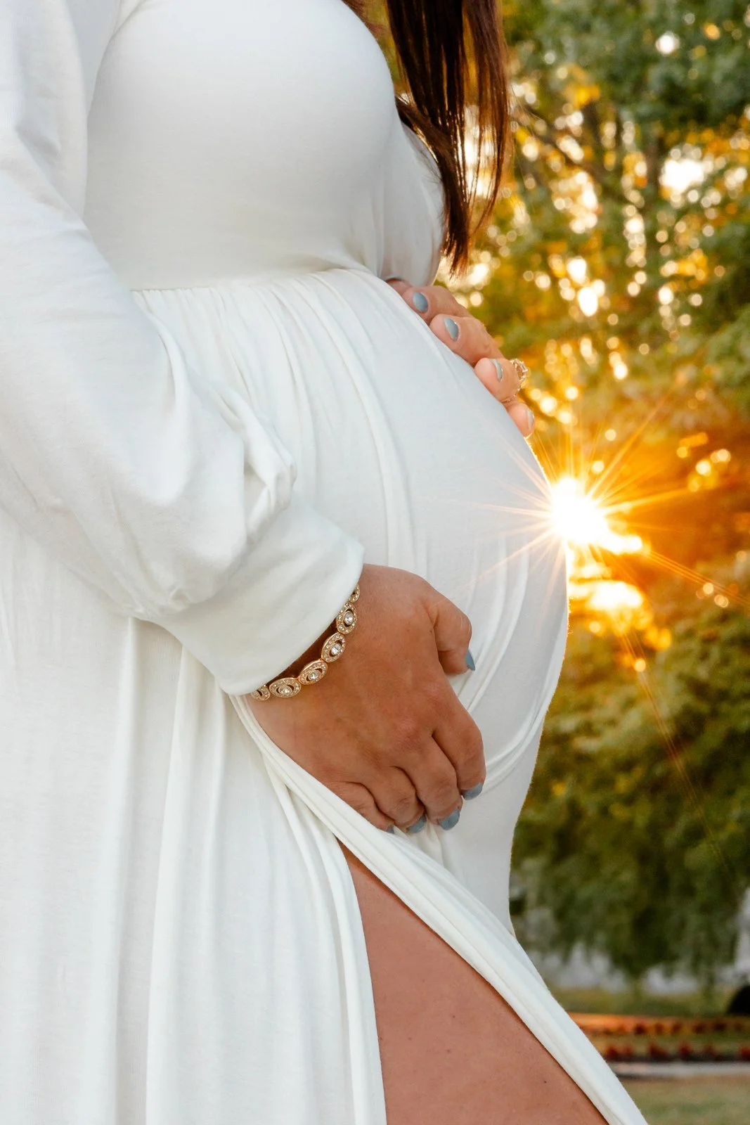 Close-up of a pregnant woman in a white dress holding her belly with both hands, standing outdoors during sunset, with the sun shining through trees in the background.