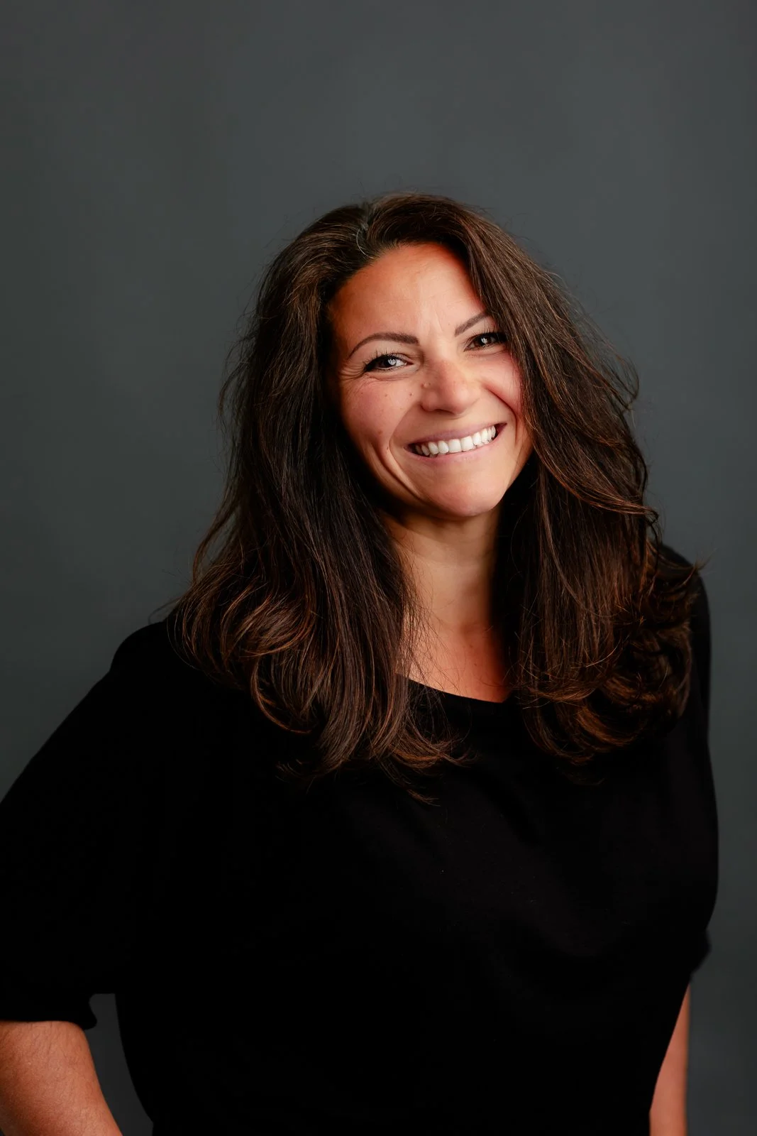 Smiling woman with long, wavy brown hair wearing a black top, standing against a dark gray background.