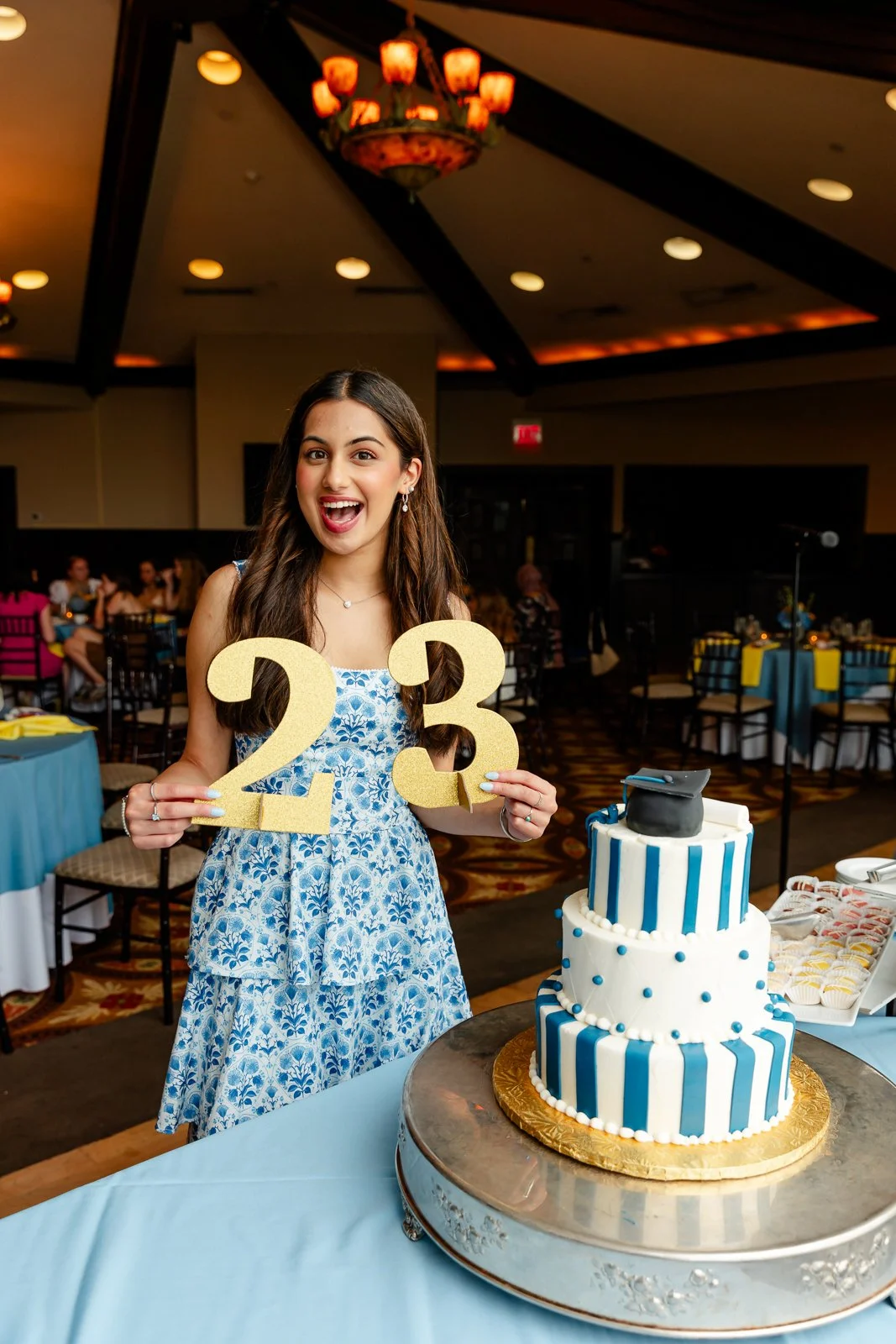 A young woman at a celebration holding large gold numbers '2' and '3' in front of a three-tiered blue and white striped cake with a graduation cap on top, in a decorated event space.