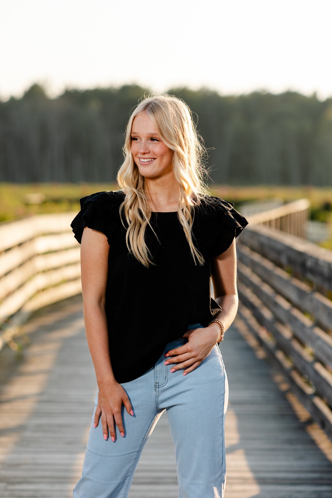 A young woman with long blonde hair wearing a black top and light blue jeans, standing on a wooden pathway outdoors during a sunny day.