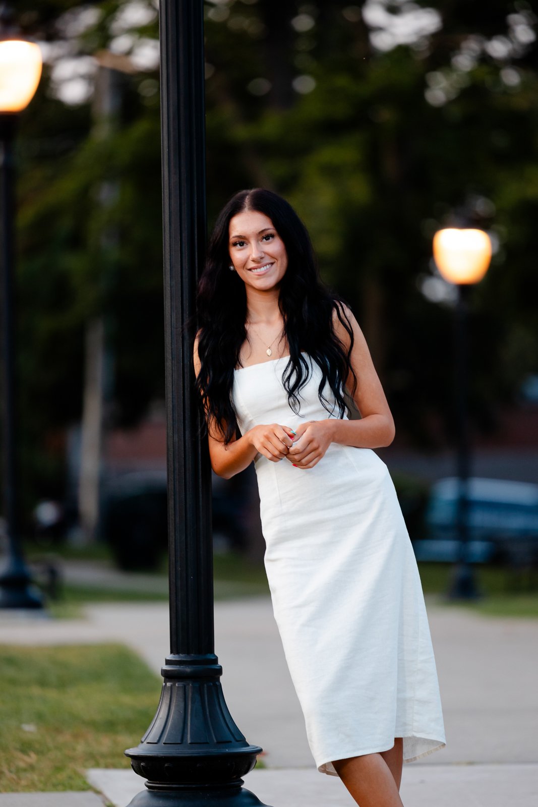 A young woman with long black hair, wearing a white sleeveless dress, smiling and leaning against a black lamppost outdoors during evening with blurred street lights and trees in the background.