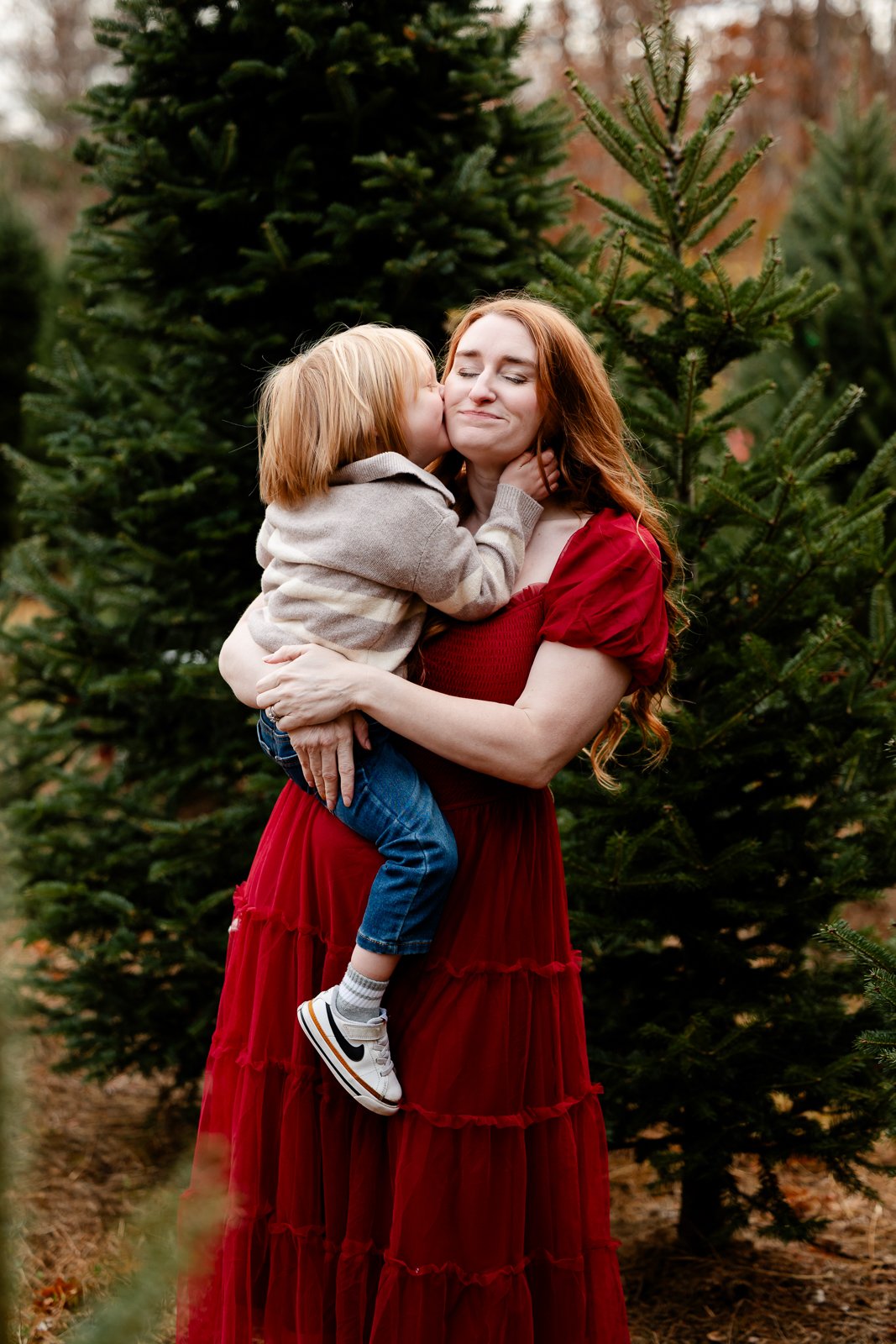 A woman in a red dress is holding a young boy, who is giving her a kiss on the cheek, surrounded by Christmas trees outdoors.