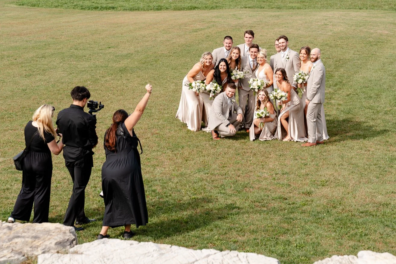 A wedding party posing on a grassy field with bridesmaids and groomsmen holding bouquets of white flowers, being photographed by a crew of three people, one of whom is waving.