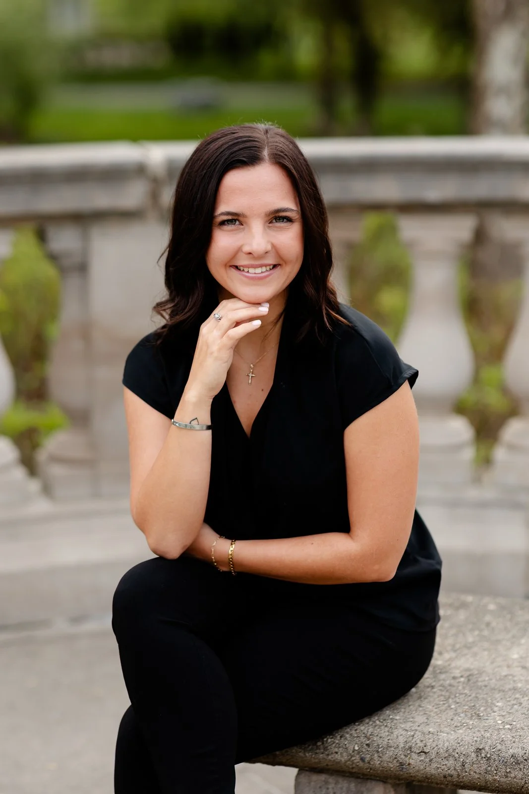 Young woman with dark hair wearing a black shirt and black pants, sitting on a stone bench outdoors in a park, smiling at the camera with her hand near her chin.