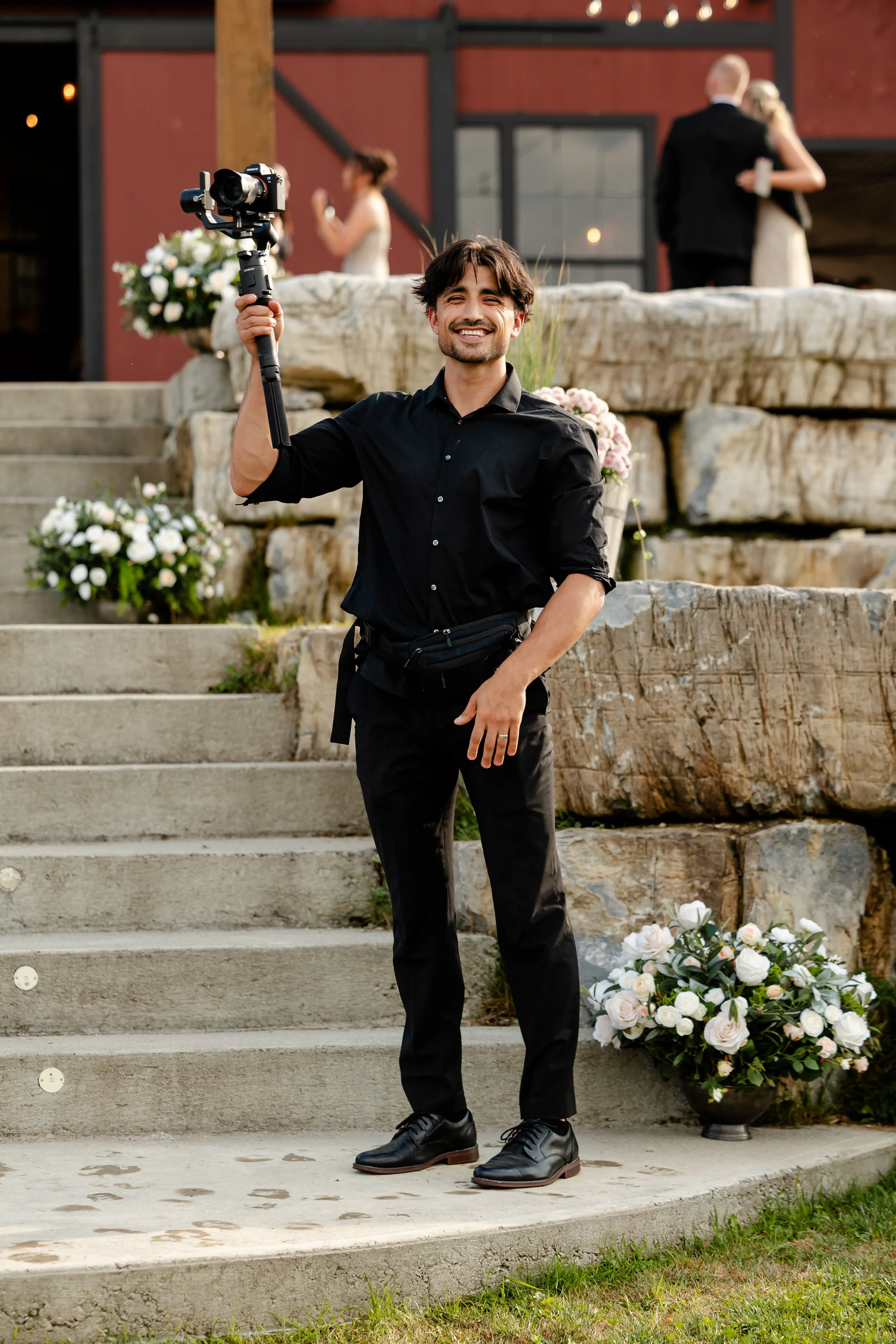 A man dressed in black holding a camera stabilizer, smiling, standing on outdoor steps decorated with white flower arrangements, with a wedding couple embracing in the background.