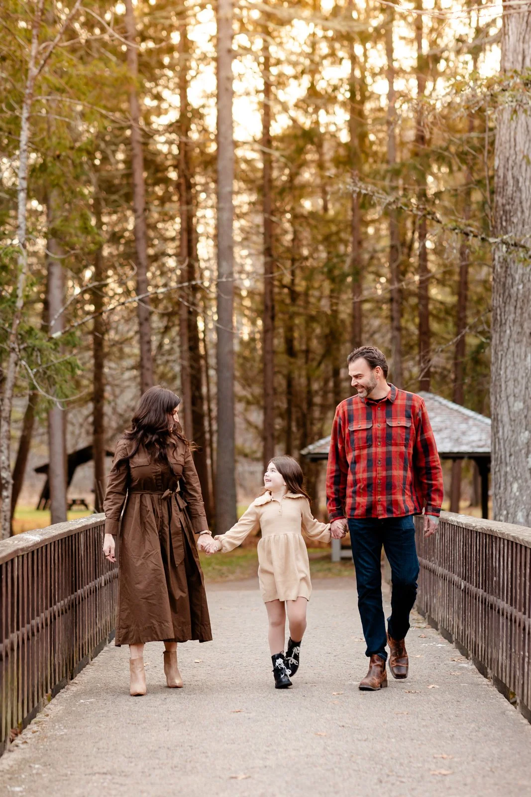 A family of three walking on a bridge in a wooded park during autumn, holding hands and smiling.