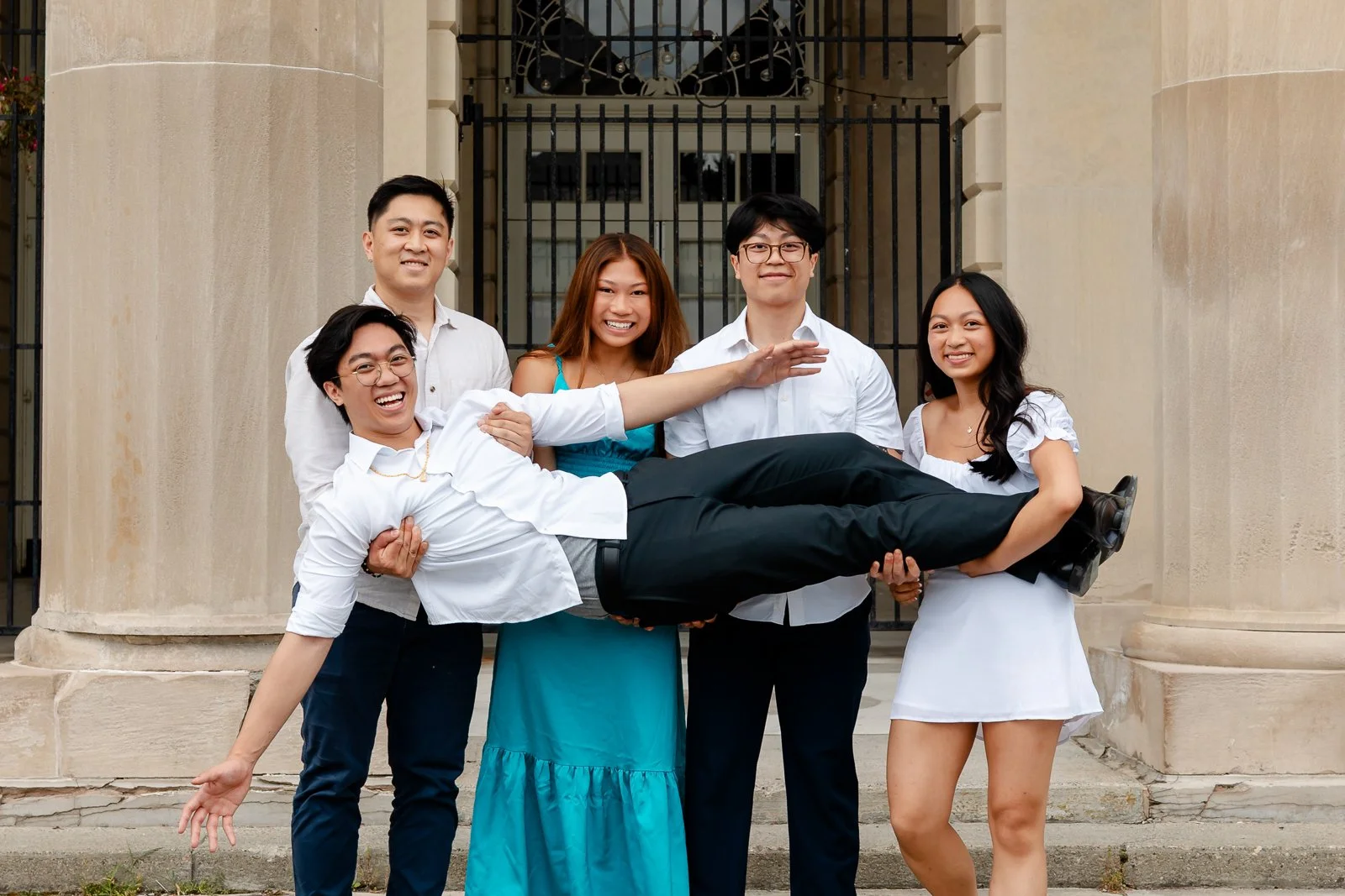 A group of five young adults standing in front of a building, with one person being held horizontally by the others. All are smiling and dressed in semi-formal attire.