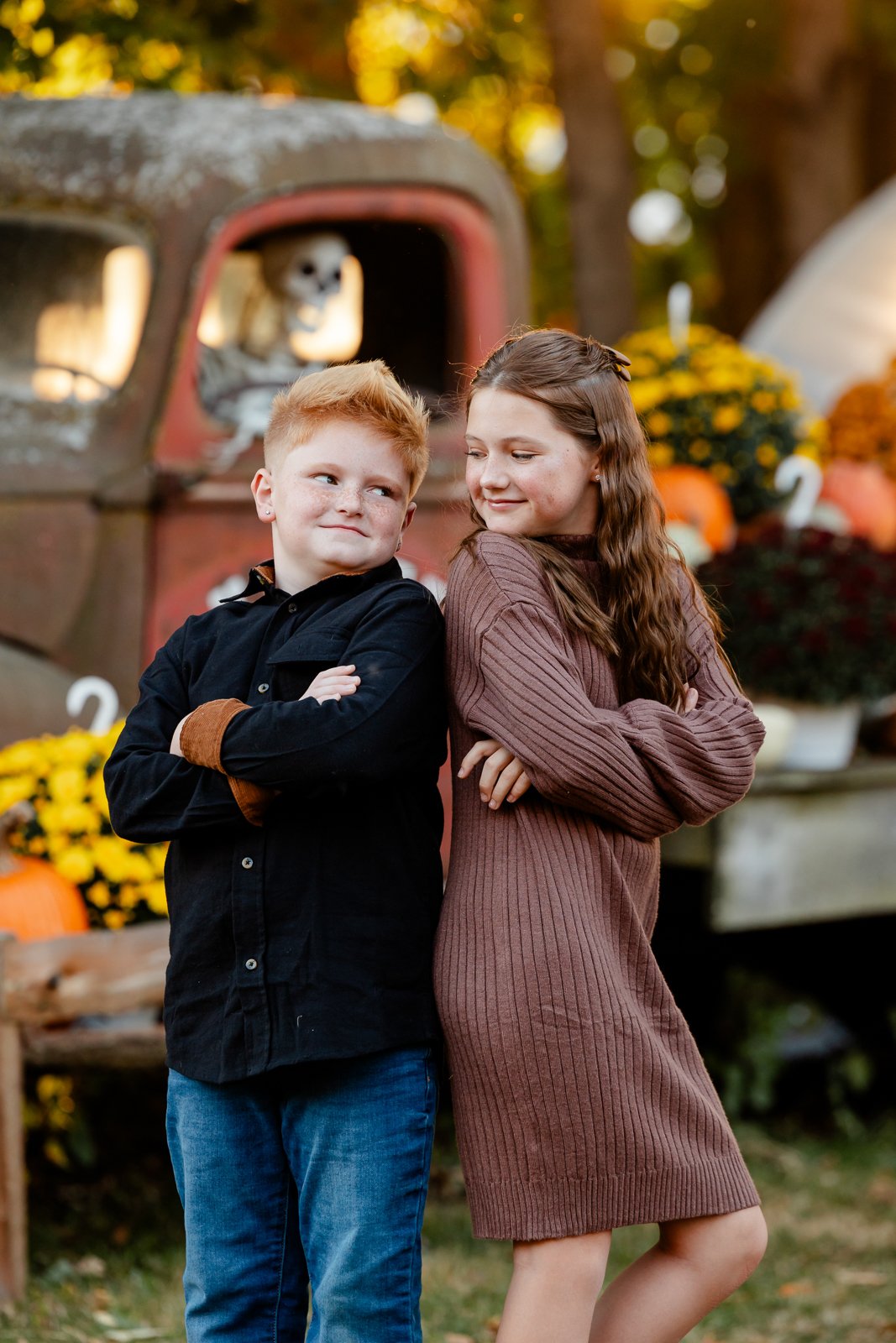 A young boy and girl standing outside with crossed arms, smiling at each other, in front of an autumn-themed backdrop featuring pumpkins, mums, and an old truck with a dog in the window.