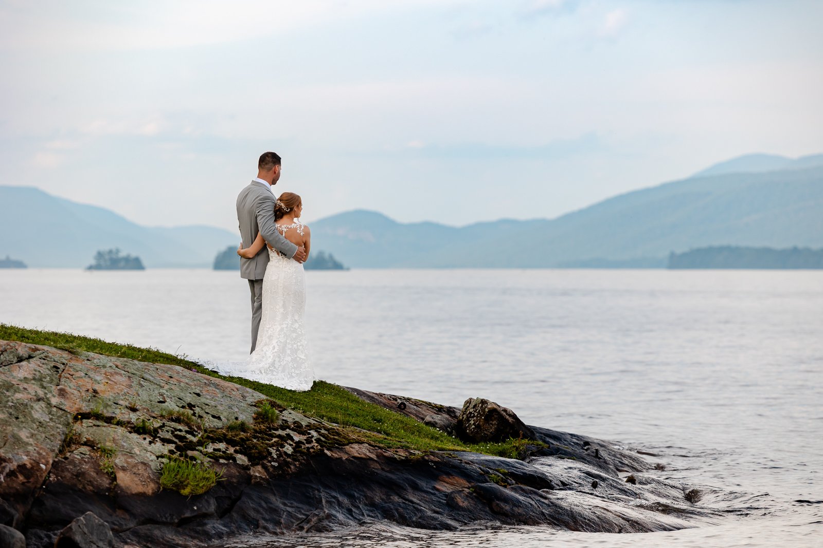 A bride and groom standing on a rocky lakeshore with mountains in the background, embracing each other.