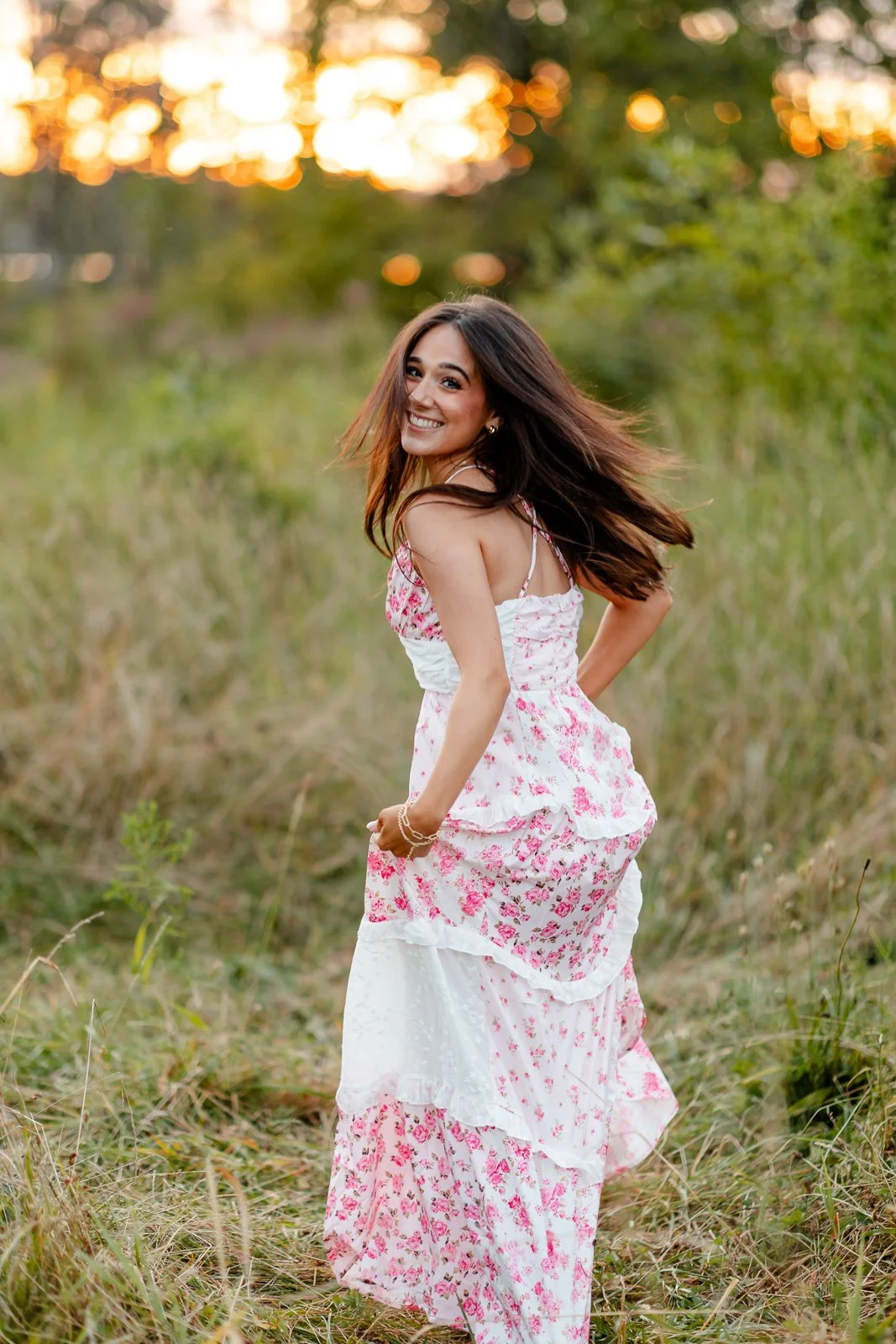 A young woman with long dark hair, wearing a white and pink floral dress, smiling and standing in a grassy field during sunset.