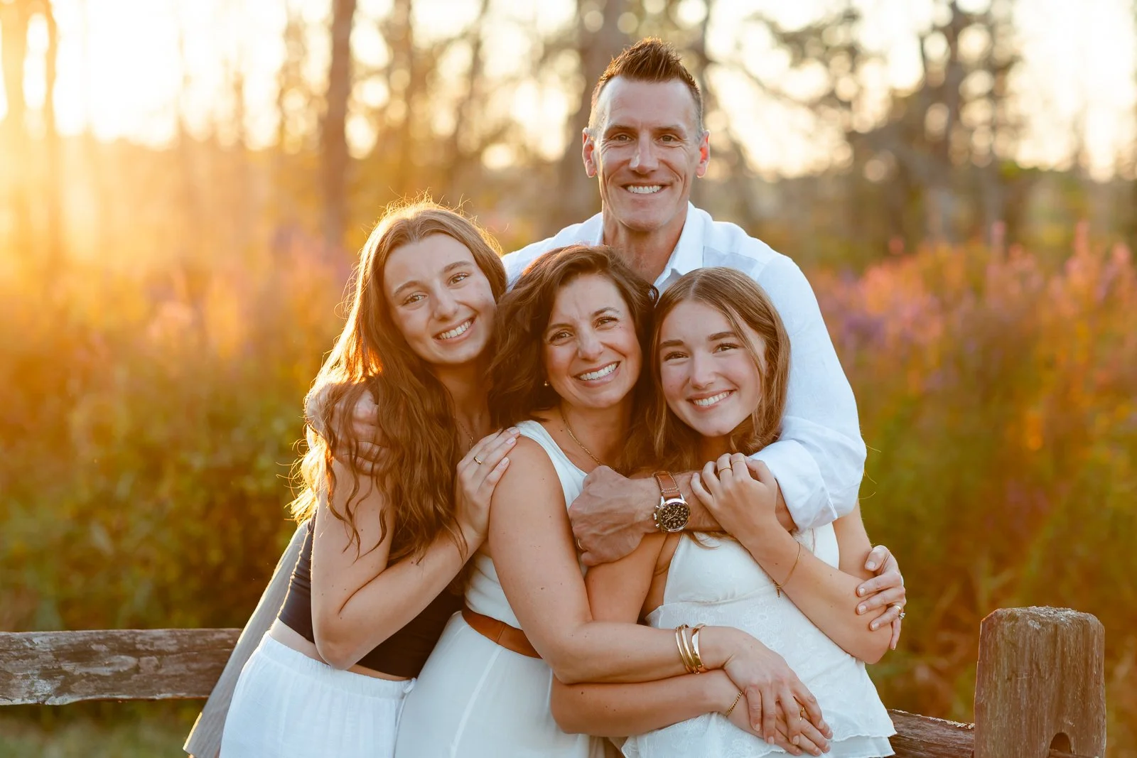 A family of four smiling and hugging outdoors during sunset, with trees and flowers in the background.