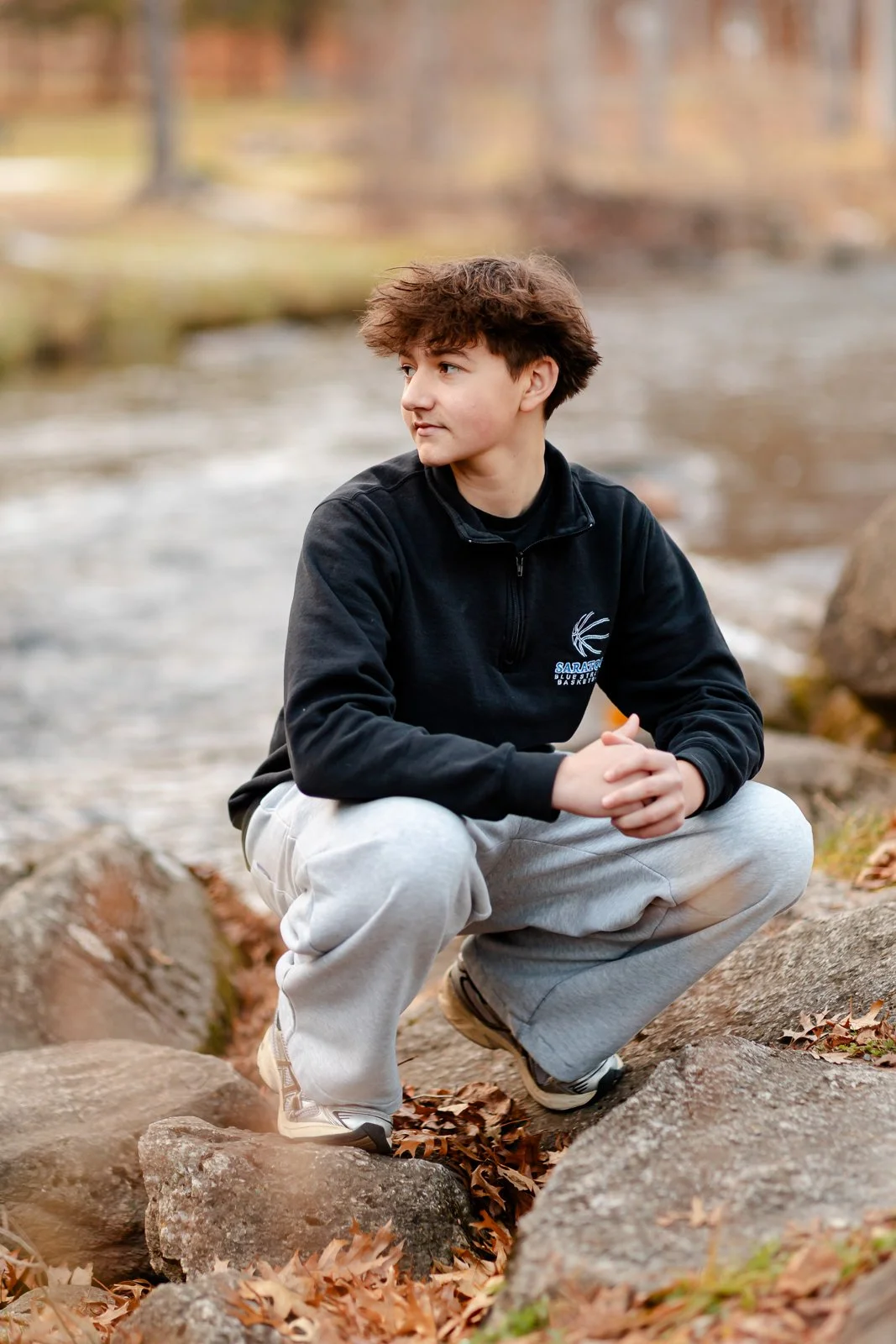 A young person squatting on rocks beside a river with autumn leaves on the ground, wearing a black sweatshirt, light gray sweatpants, and sneakers, looking to the side.