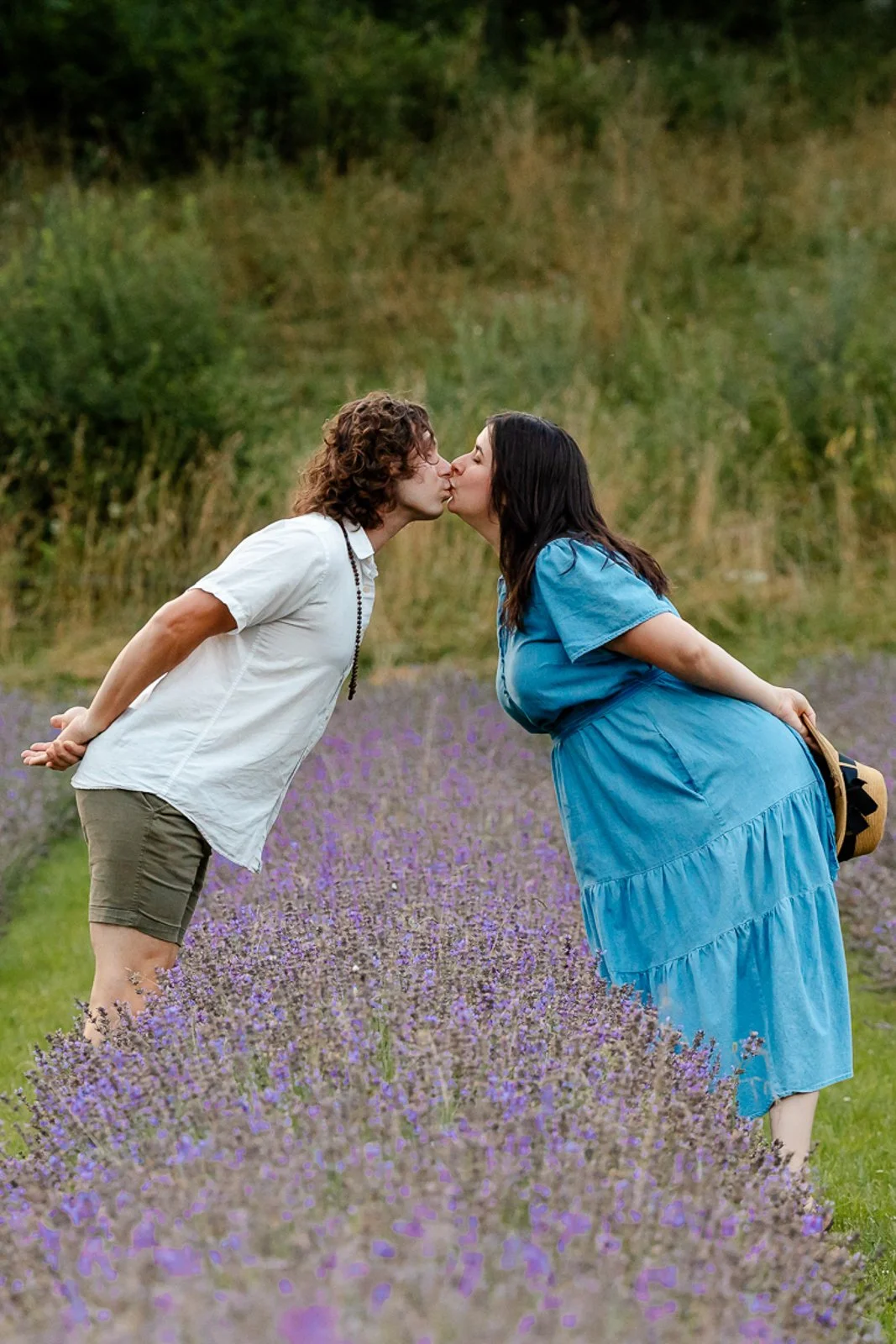 A man and woman kiss in a lavender field, with the woman wearing a blue dress and the man wearing a white shirt and shorts.