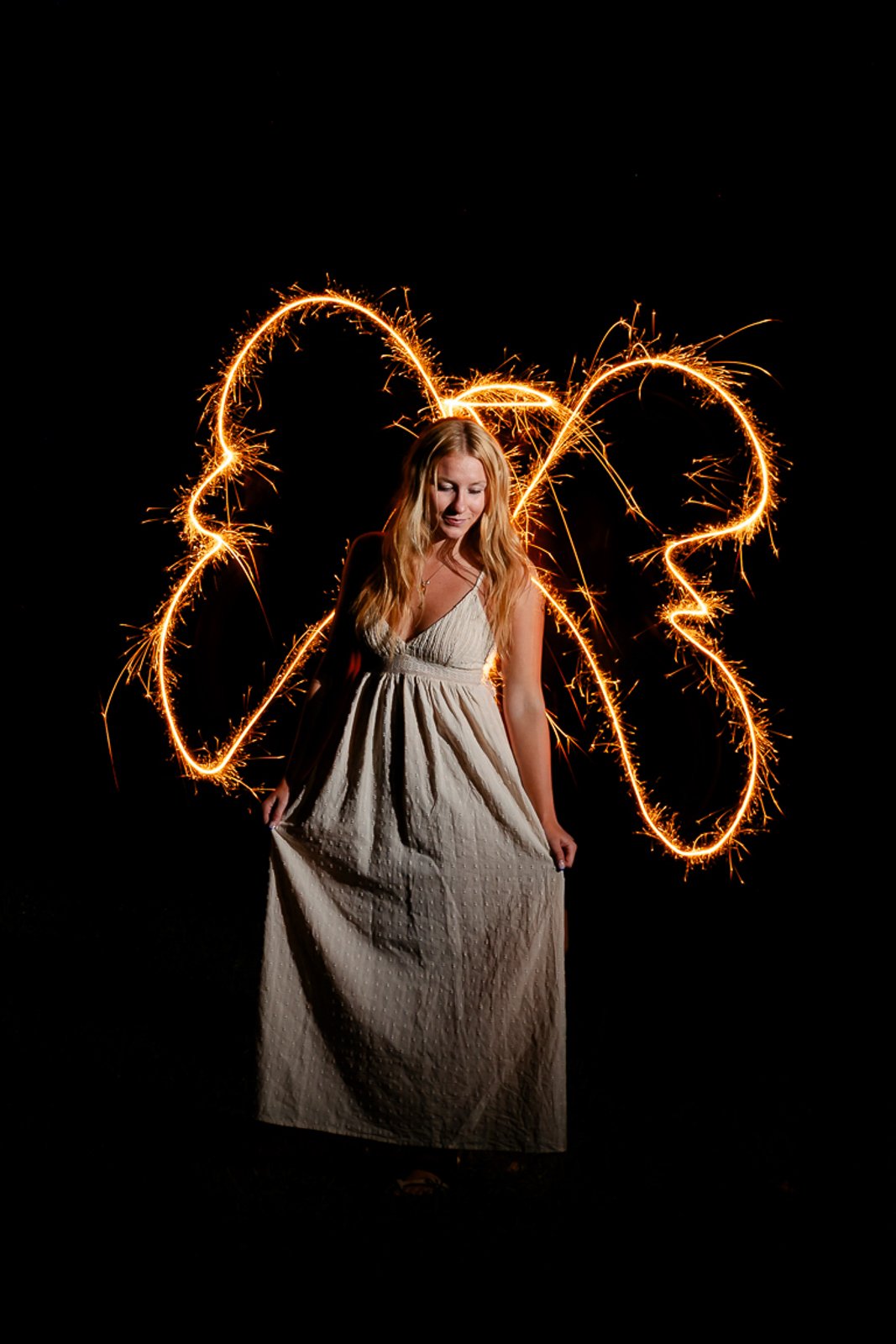 A young woman in a white dress standing in the dark with sparkler light trails forming butterfly wings behind her.