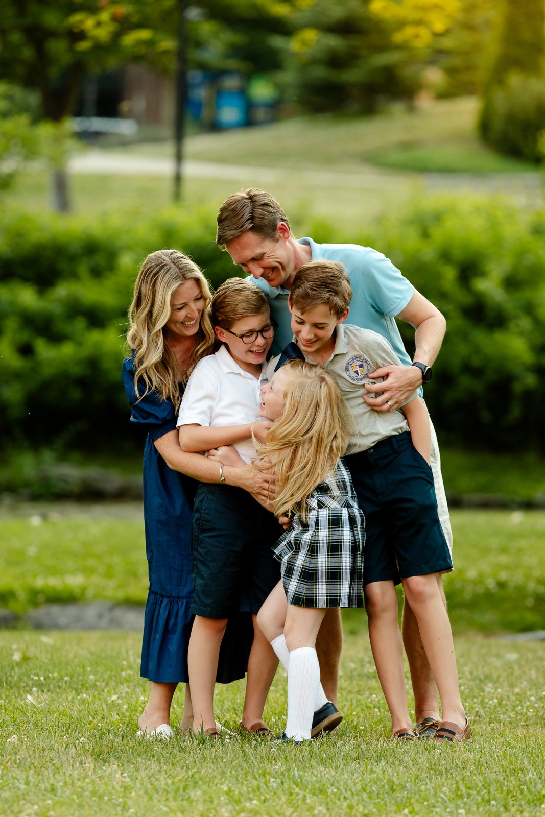 A happy family of six, including children and adults, laughing and hugging outdoors on a grassy area in a park.