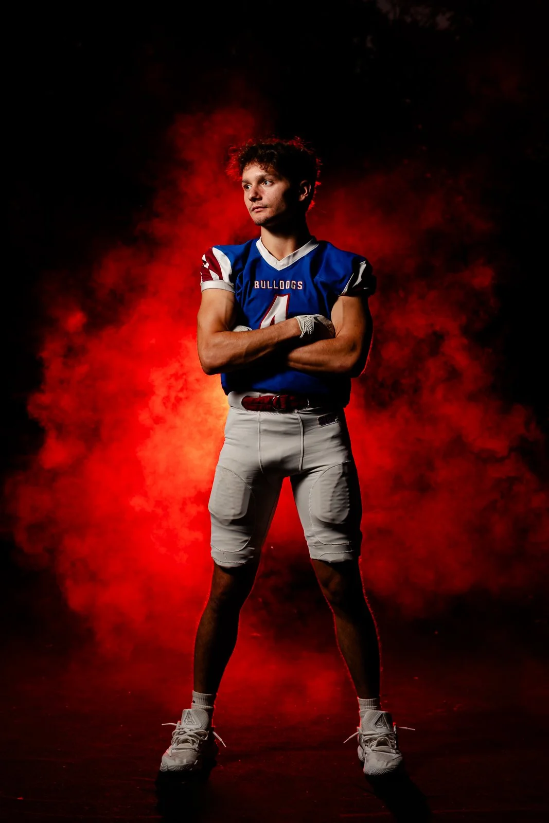 A young male football player in a blue and white uniform with the word 'Bulldogs' on the chest, standing with arms crossed in front of a red smoky background.