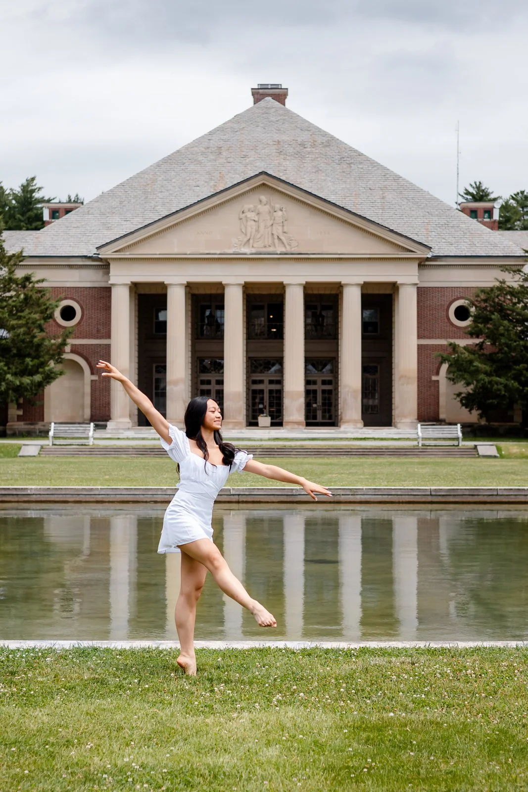 A woman in a white dress dancing on grass beside a pond with a historic building in the background.