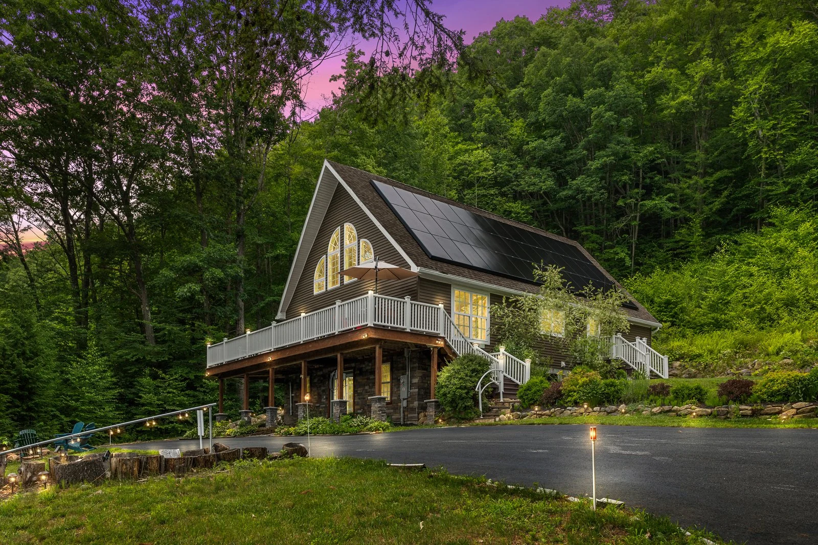 A house with a steep roof covered in solar panels, large arched windows, a wooden deck with an umbrella, surrounded by greenery at dusk with a the sky showing purple hues.