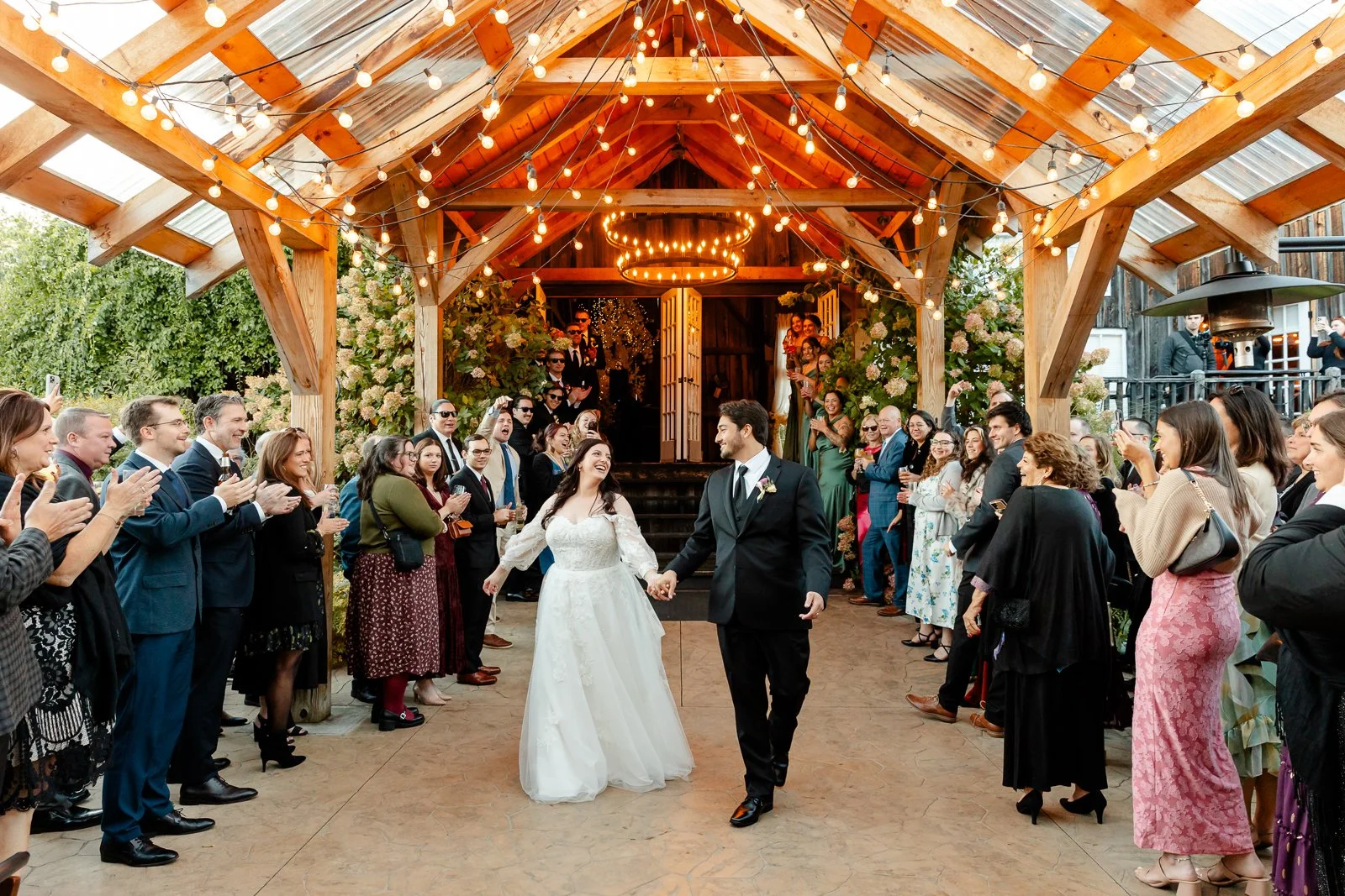 A bride and groom holding hands, walking through a crowd of guests at a wedding reception under a decorated wooden pavilion with string lights, with guests clapping and smiling.