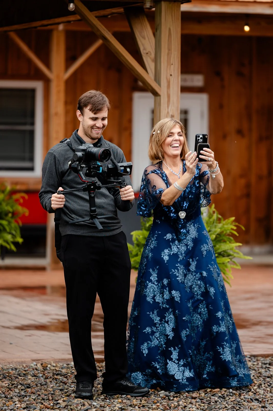 A woman in a blue floral dress and a man holding a camera are smiling and looking at a phone, standing outdoors on a pebble ground with green plants and a wooden building in the background.