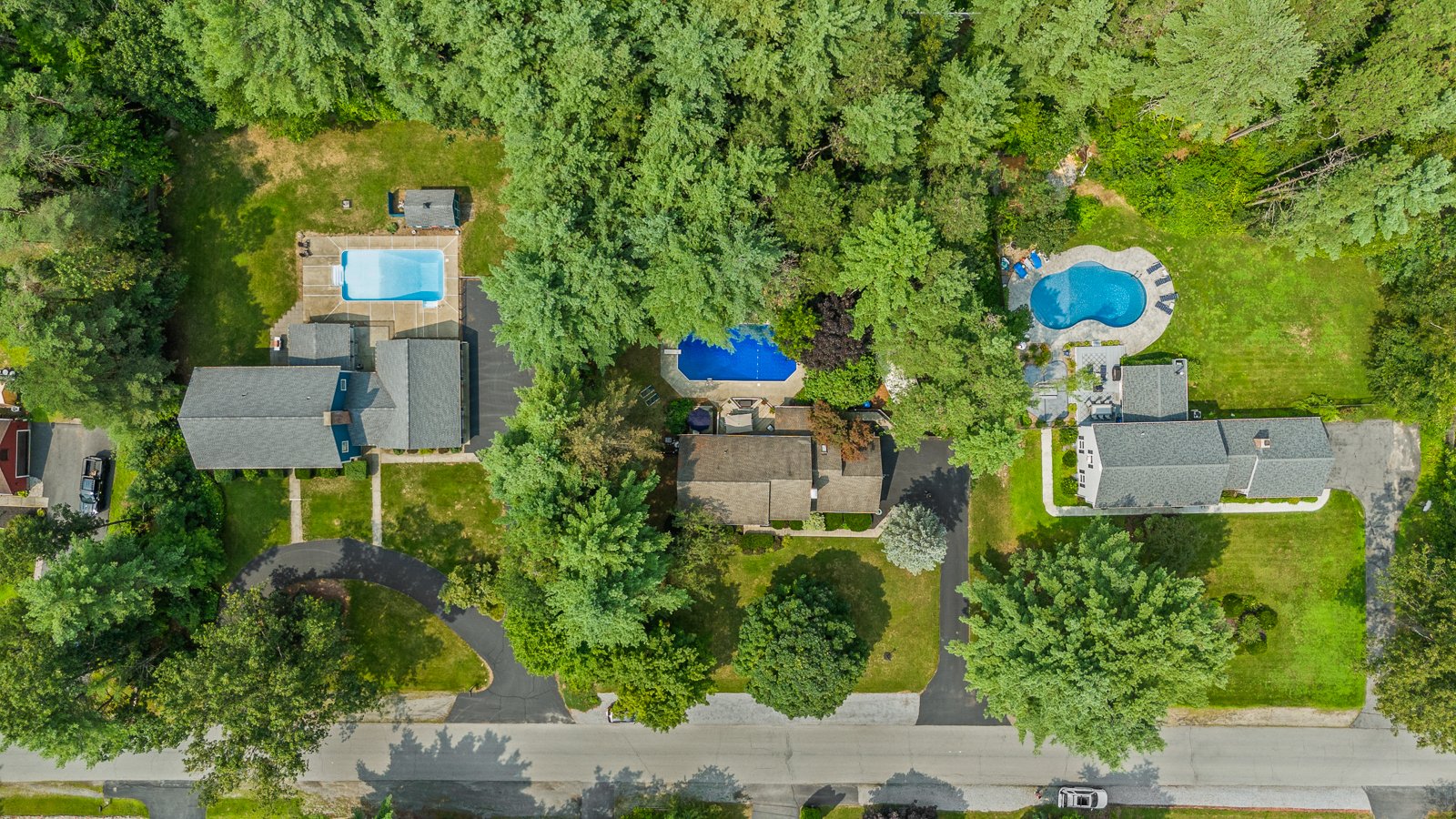 Aerial view of several houses with yards and swimming pools, surrounded by trees.