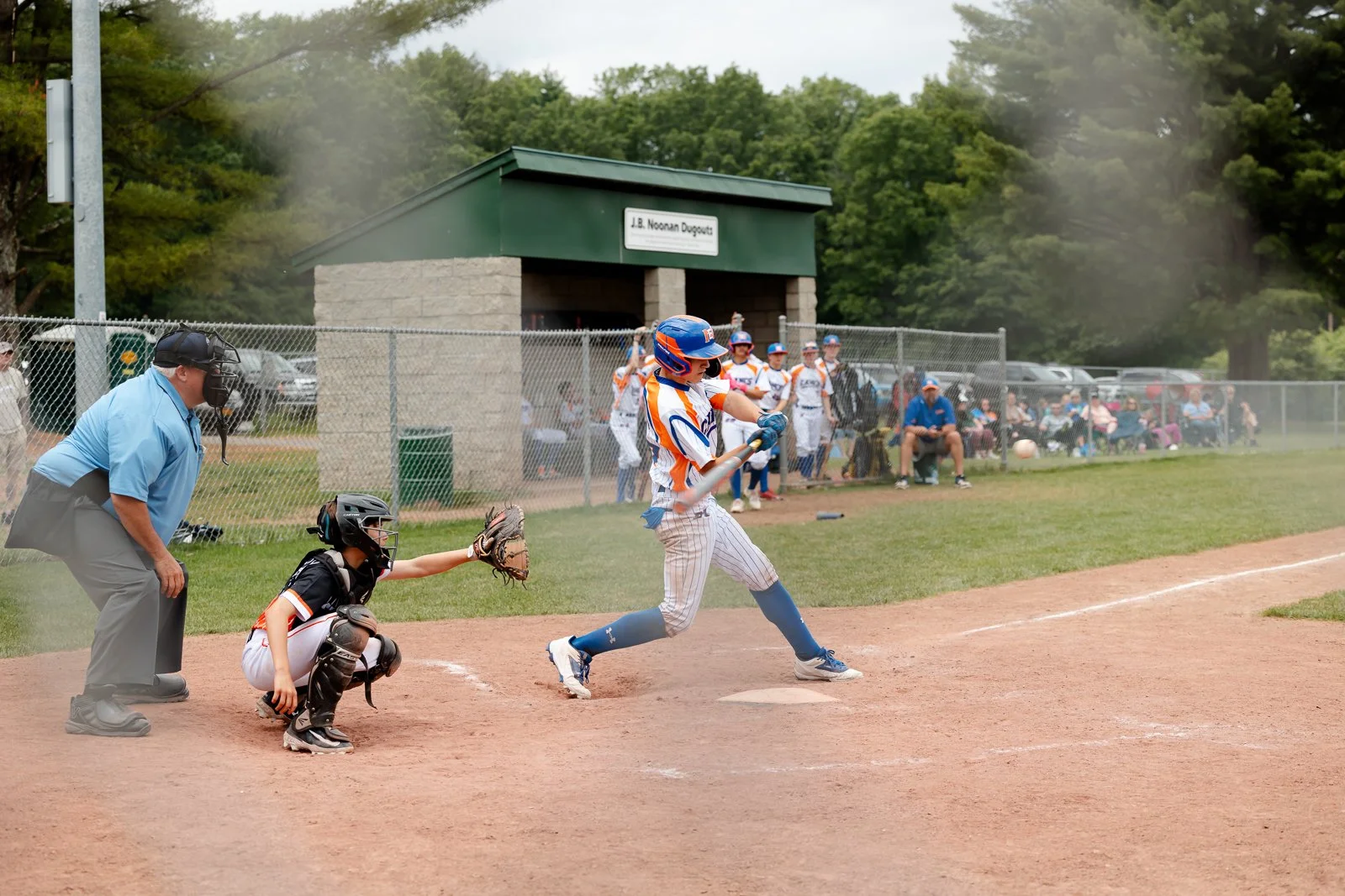 A young baseball player is swinging a bat at a pitch during a game, with a catcher and umpire behind him, and spectators watching from the stands and bench area in the background.