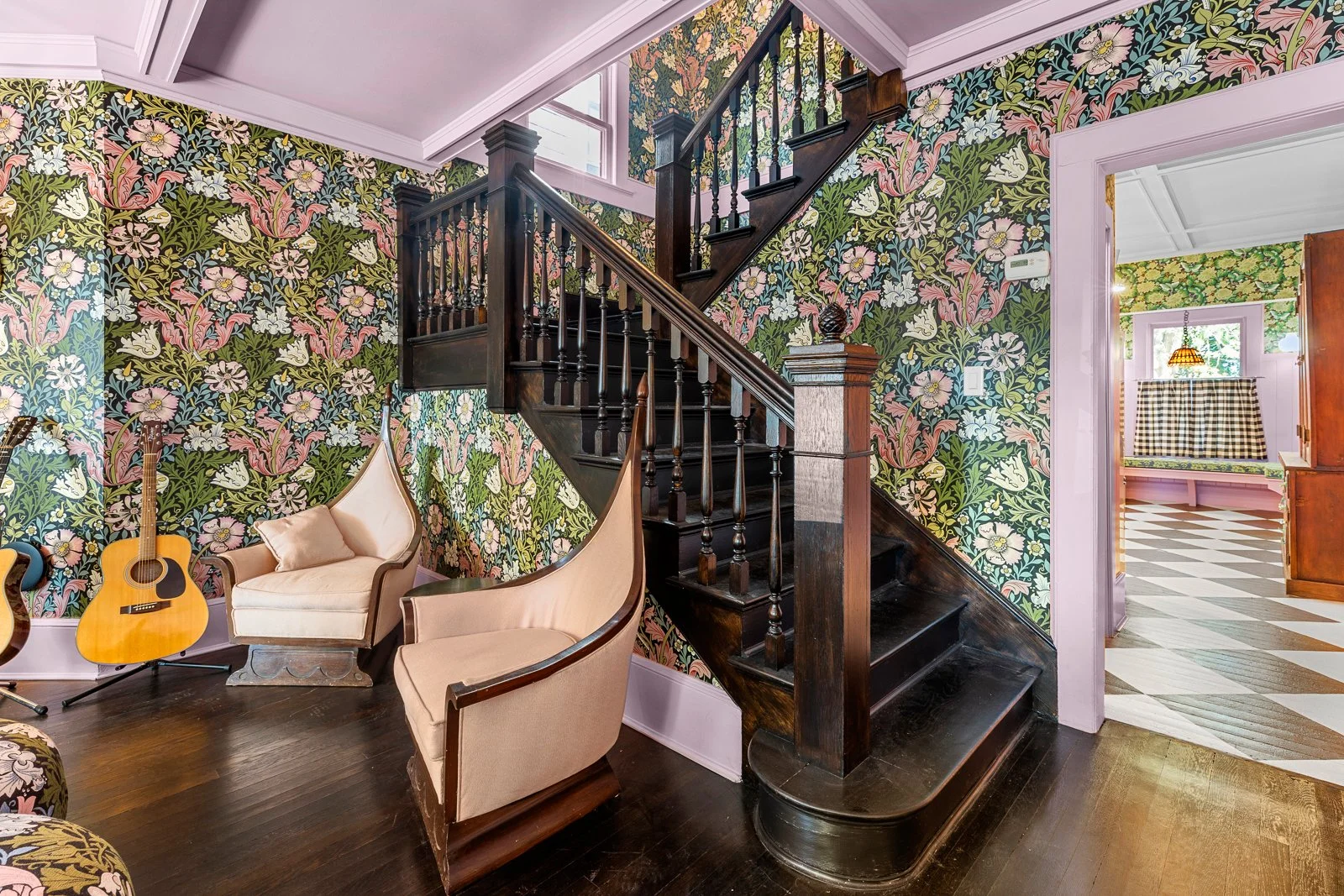 A staircase with dark wood railings and balusters in a room with floral wallpaper. There is a cream-colored armchair and an acoustic guitar next to it. A doorway reveals another room with pink walls and a checkered window curtain.