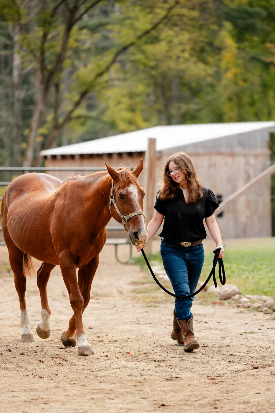 A woman walking a horse on a leash outdoors at a farm or stable, with trees and a building in the background.