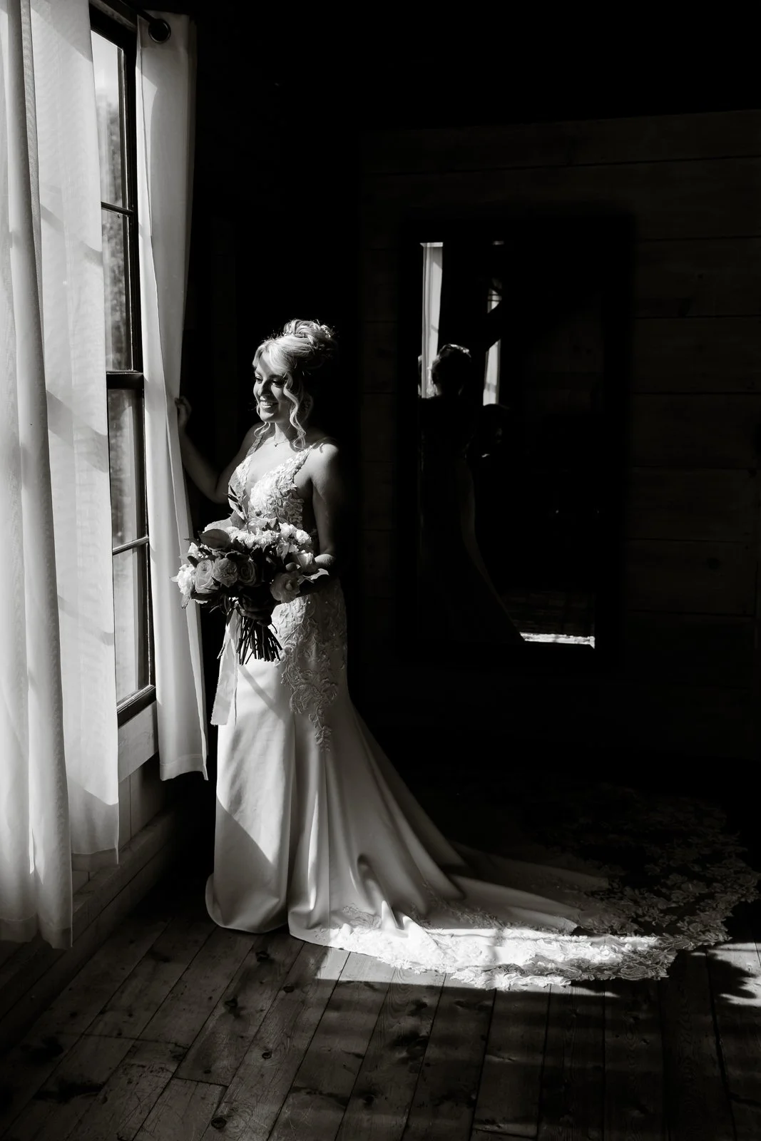 Bride in a lace wedding dress holding a bouquet, standing by a window with light coming through, in a dimly lit room with wooden walls and a mirror reflecting her silhouette.