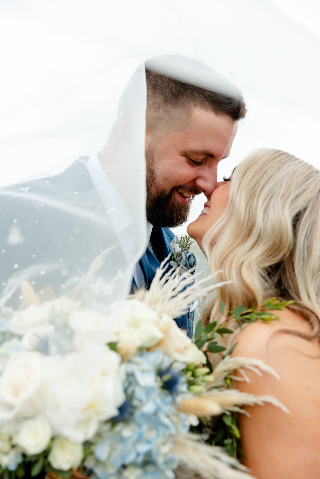 Close-up of a bride and groom smiling, with their foreheads touching, under a transparent veil, holding a bridal bouquet with white and blue flowers and greenery.